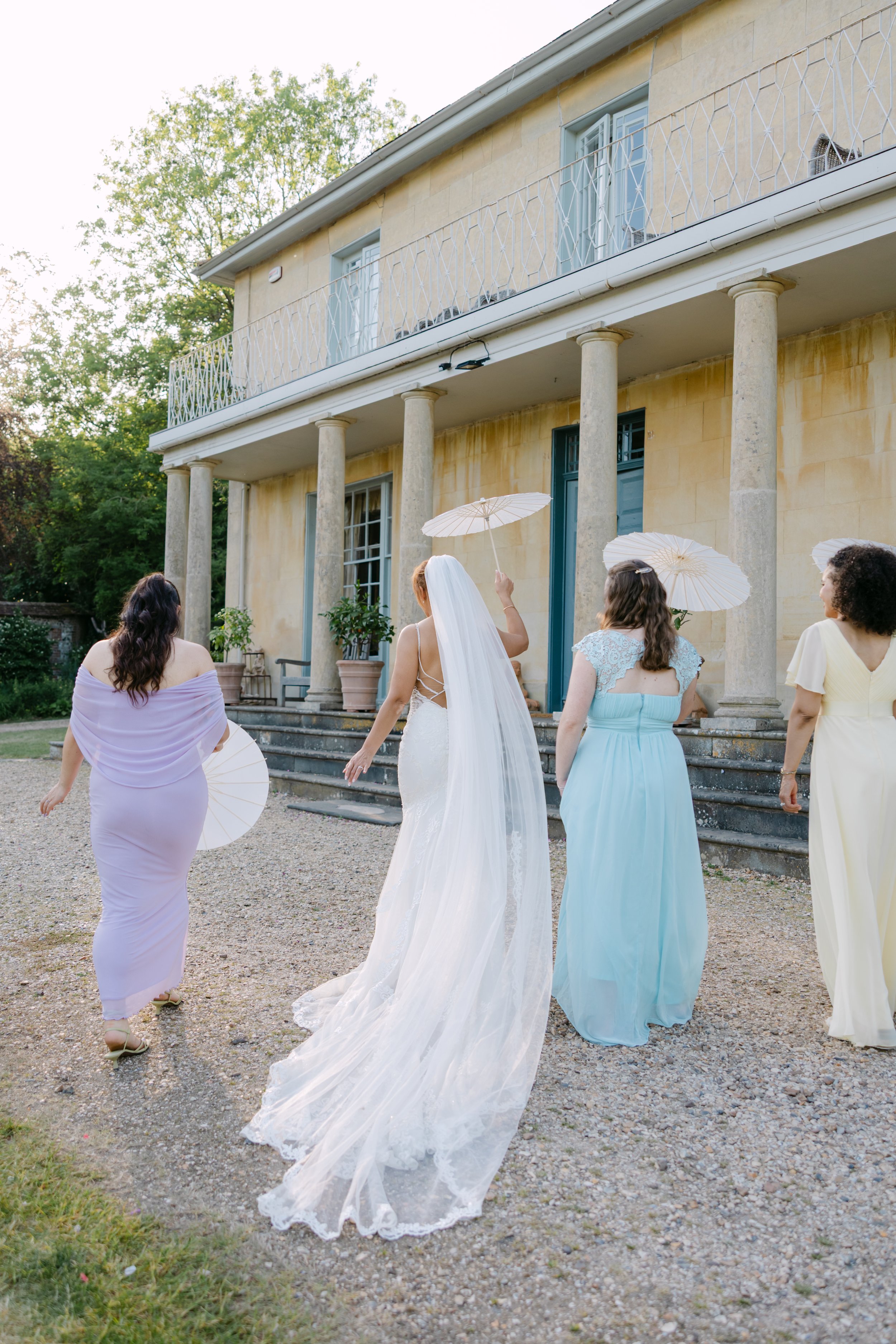 Bride walks away from camera with her bridesmaids who are dressed in full length dresses in pastel colours