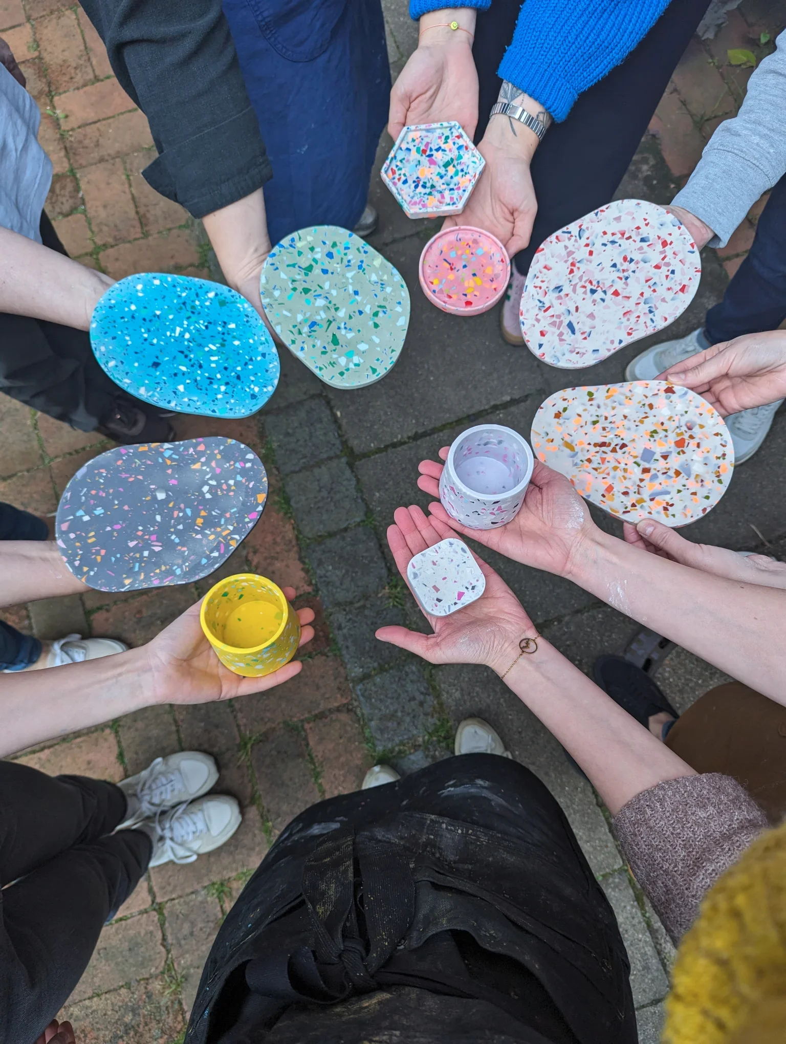 Mehrere Menschen halten bunte, marmorierte Keramikgefäße und -platten in den Händen, während sie auf einer gepflasterten Fläche stehen.