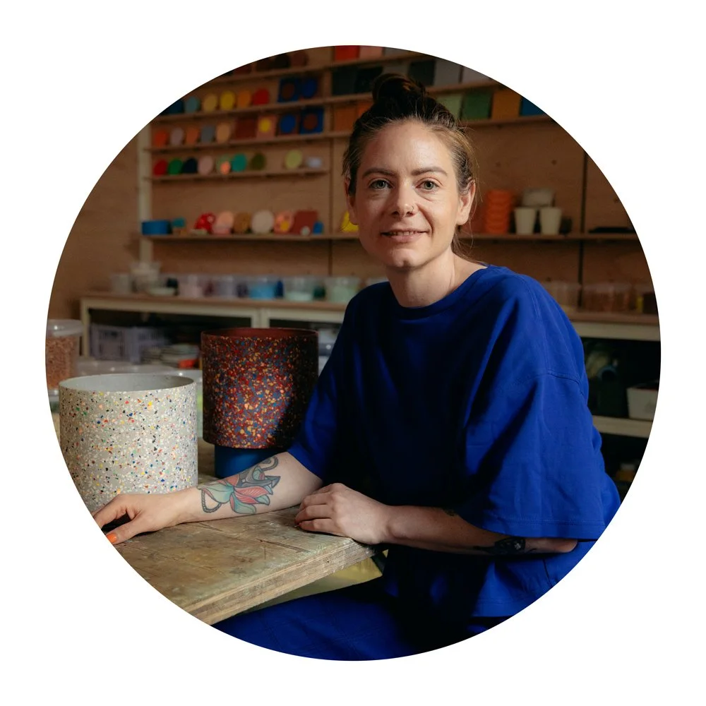 Frau sitzt an einem Tisch in einer Werkstatt mit bunten, mit Spritzmustern verzierten Keramik- oder Tongefäßen.