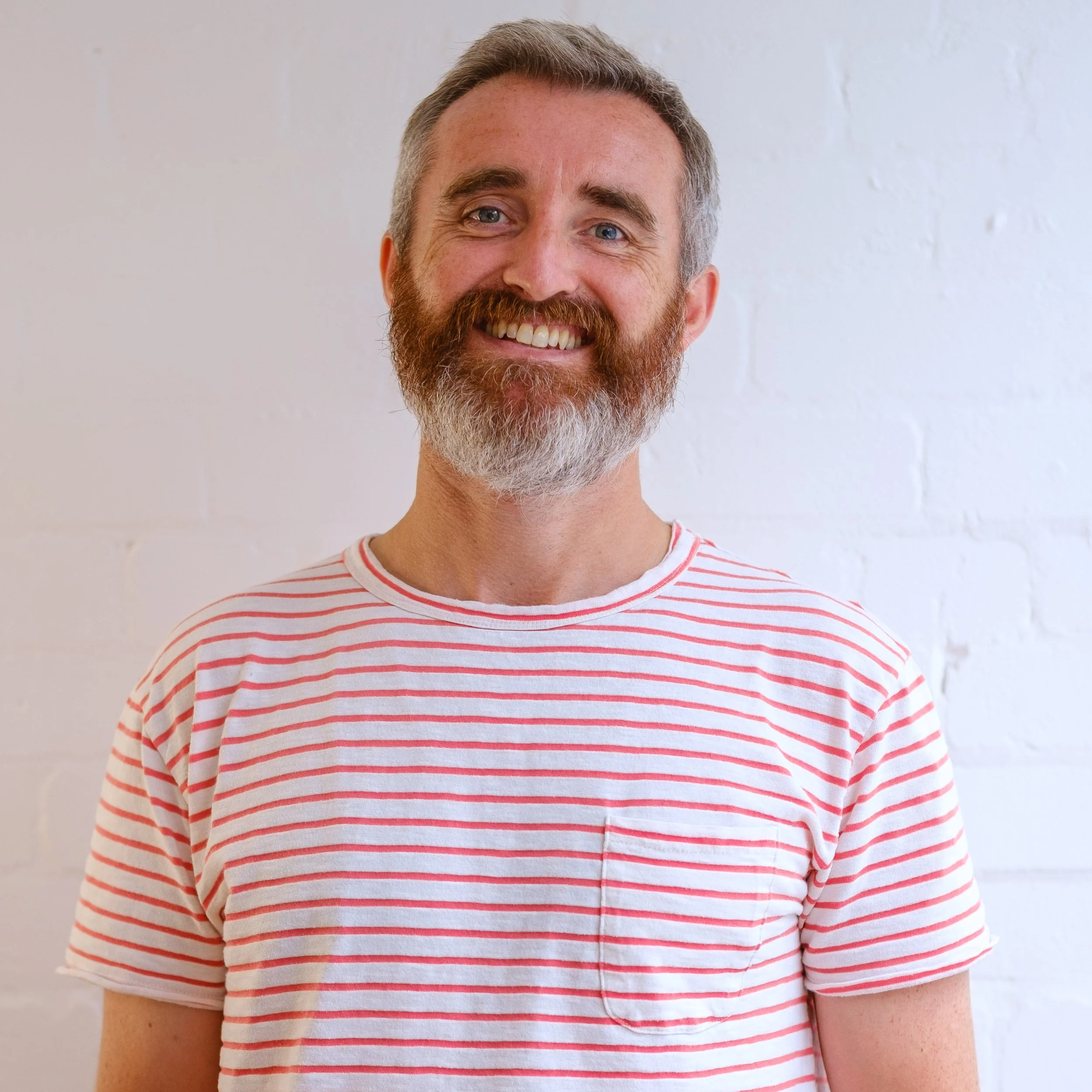 A smiling man with a full beard and short gray hair, wearing a white and red striped t-shirt, standing against a white brick wall.