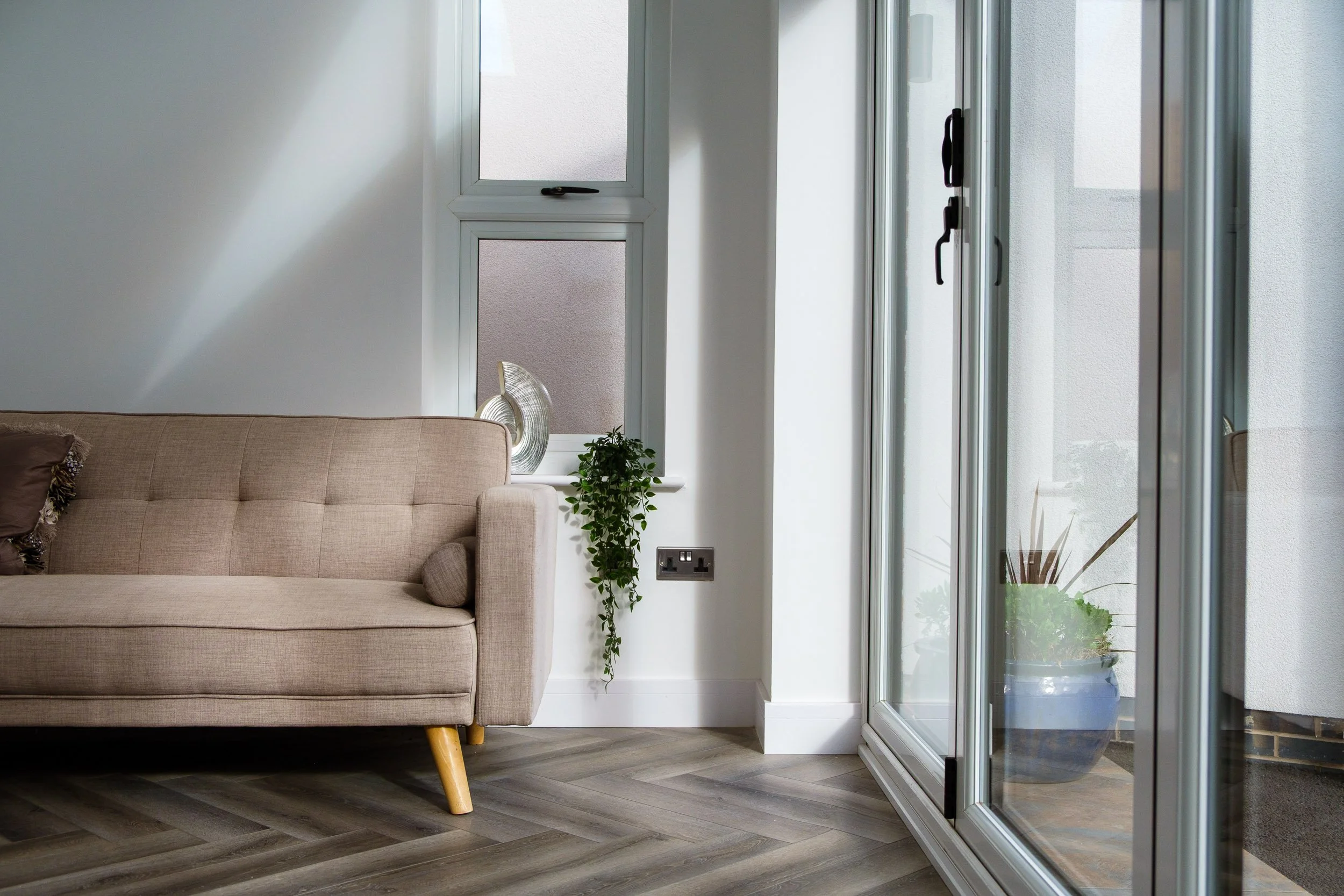 Living room with beige sofa, wooden floor, potted plants on window sill and outside balcony, and a large glass door