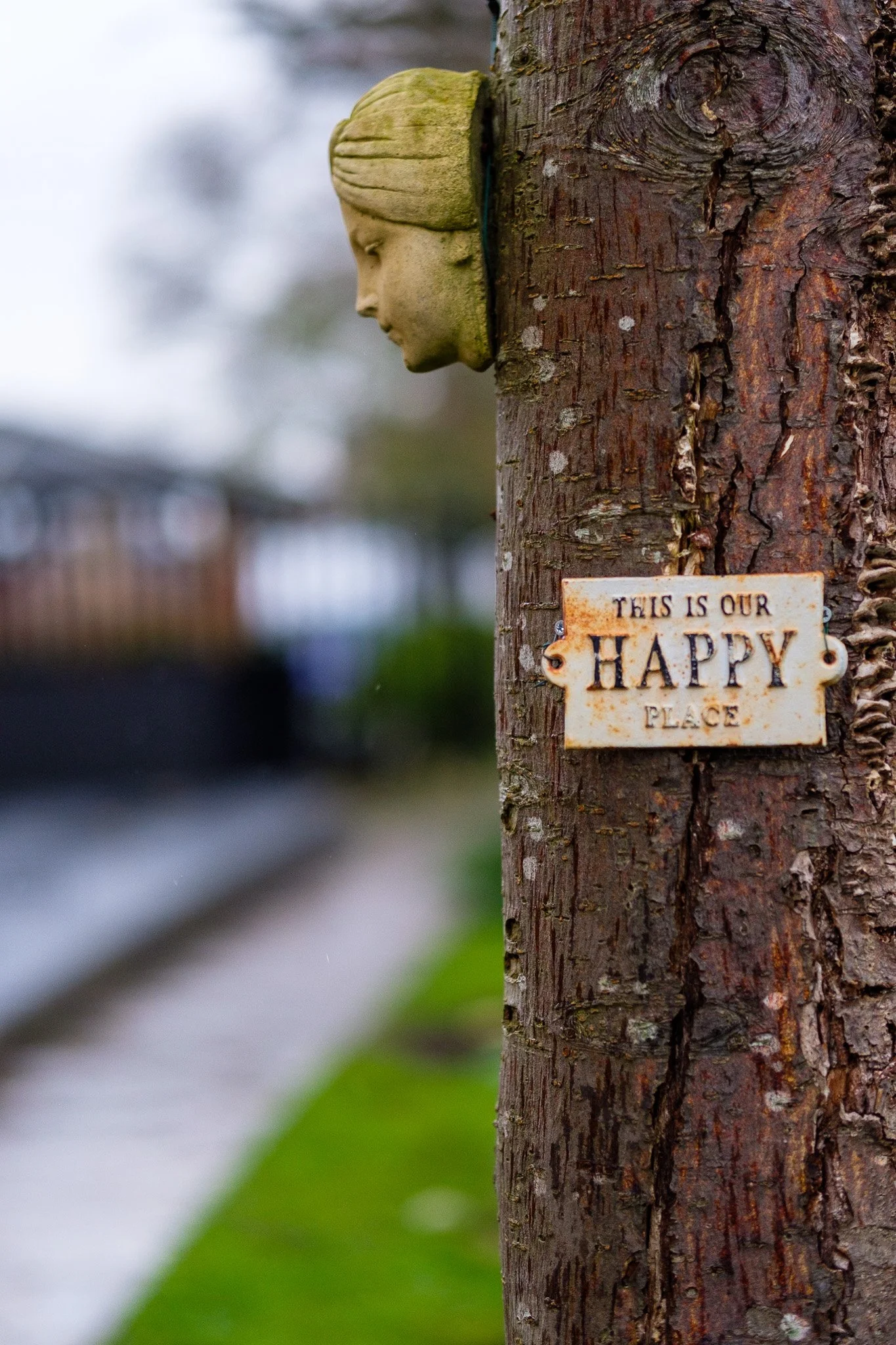 A wooden utility pole with a decorative green face sculpture of a girl's profile mounted on it. There is a rusted metal sign attached to the pole that reads, 'This is our happy place.' Blurred greenery and a railway track are visible in the backgroun