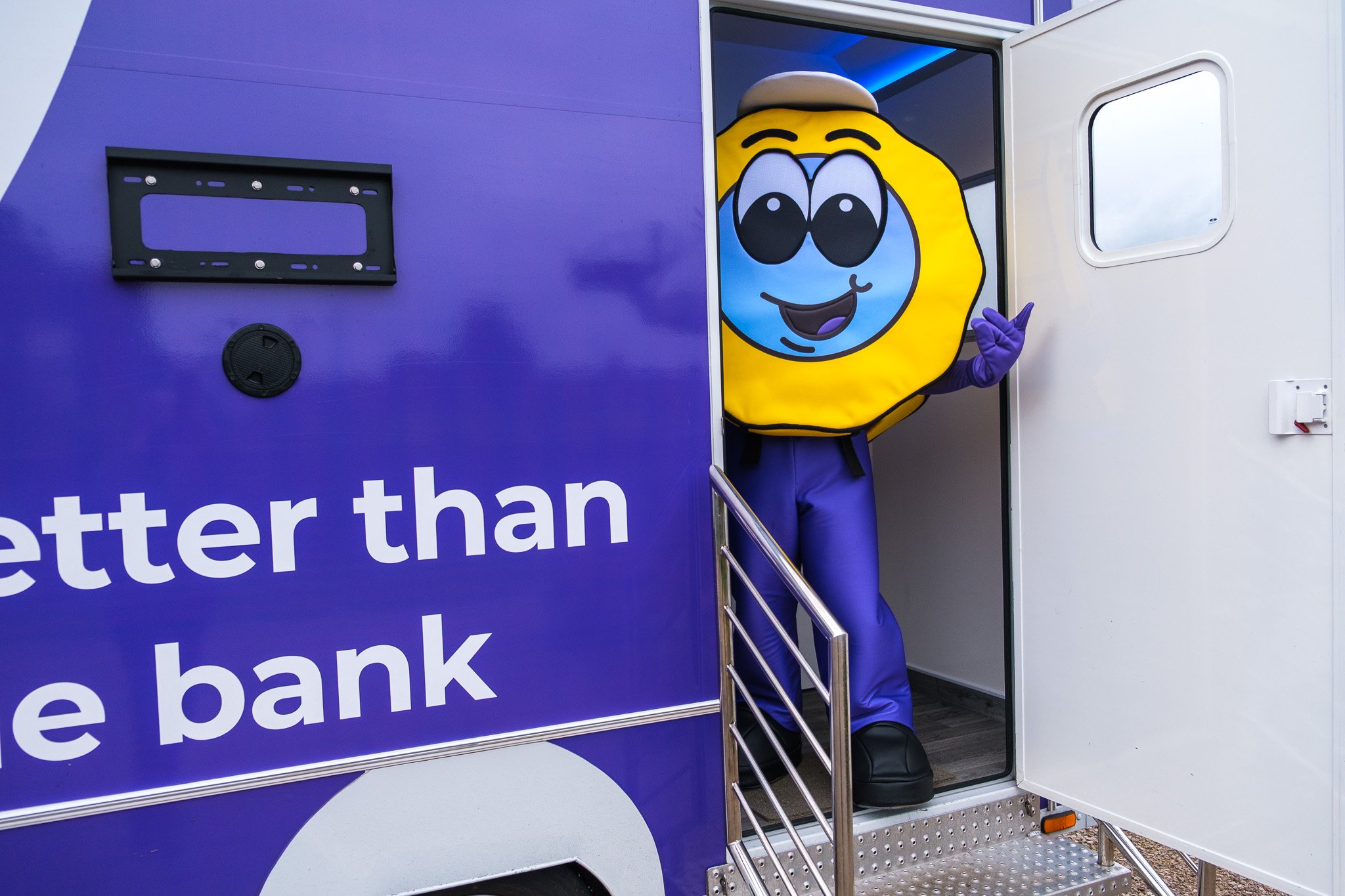 A person dressed in a yellow and blue mascot costume with large eyes and a smile, waving from the doorway of a purple and white mobile bank truck.