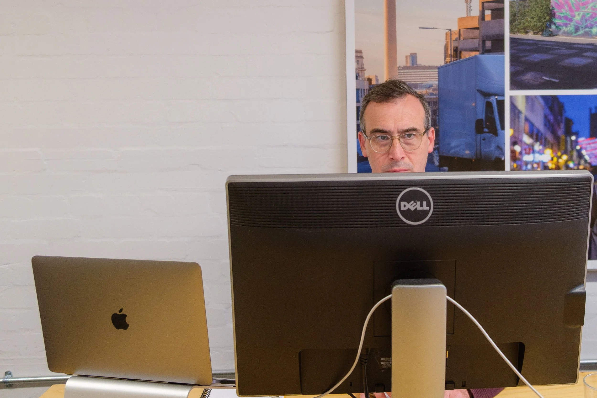 Man with glasses working at a desk, looking at a Dell computer monitor with a silver MacBook laptop beside him, against a white brick wall with cityscape photos.