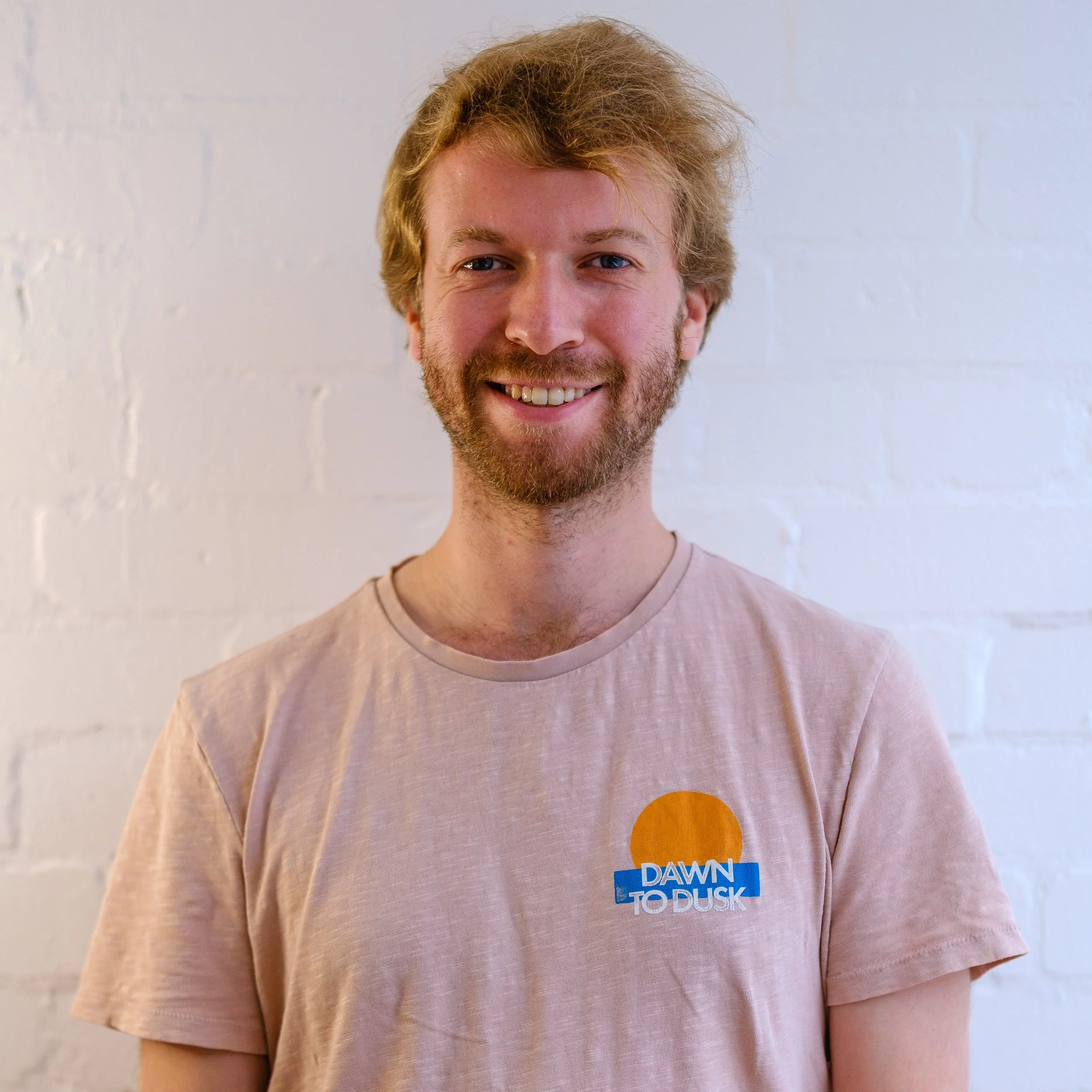 A smiling man with light brown hair, a beard, and blue eyes, wearing a light pink T-shirt with a graphic of a sun and the words 'Dawn to Dusk,' standing in front of a white brick wall.