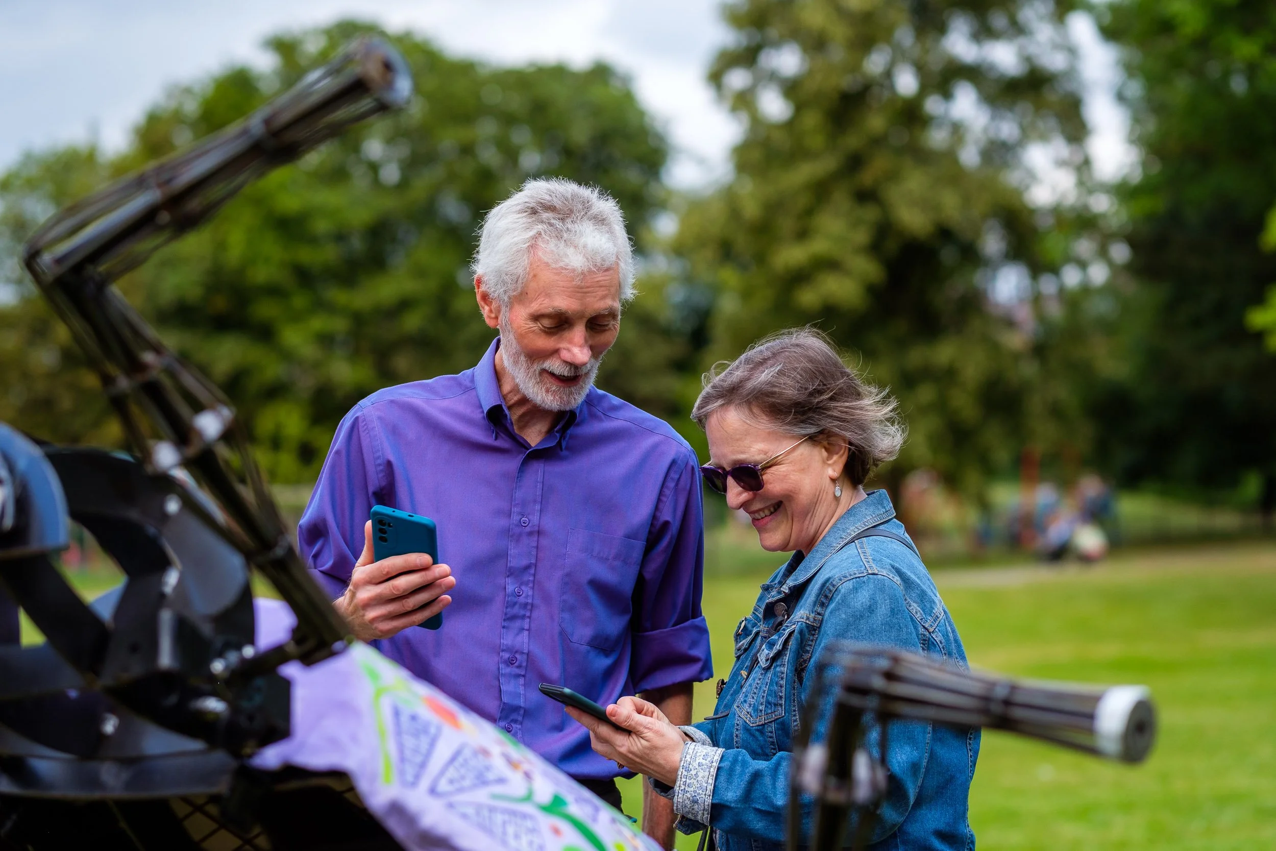 An older man and woman standing outdoors in a park, both looking at their smartphones and smiling.