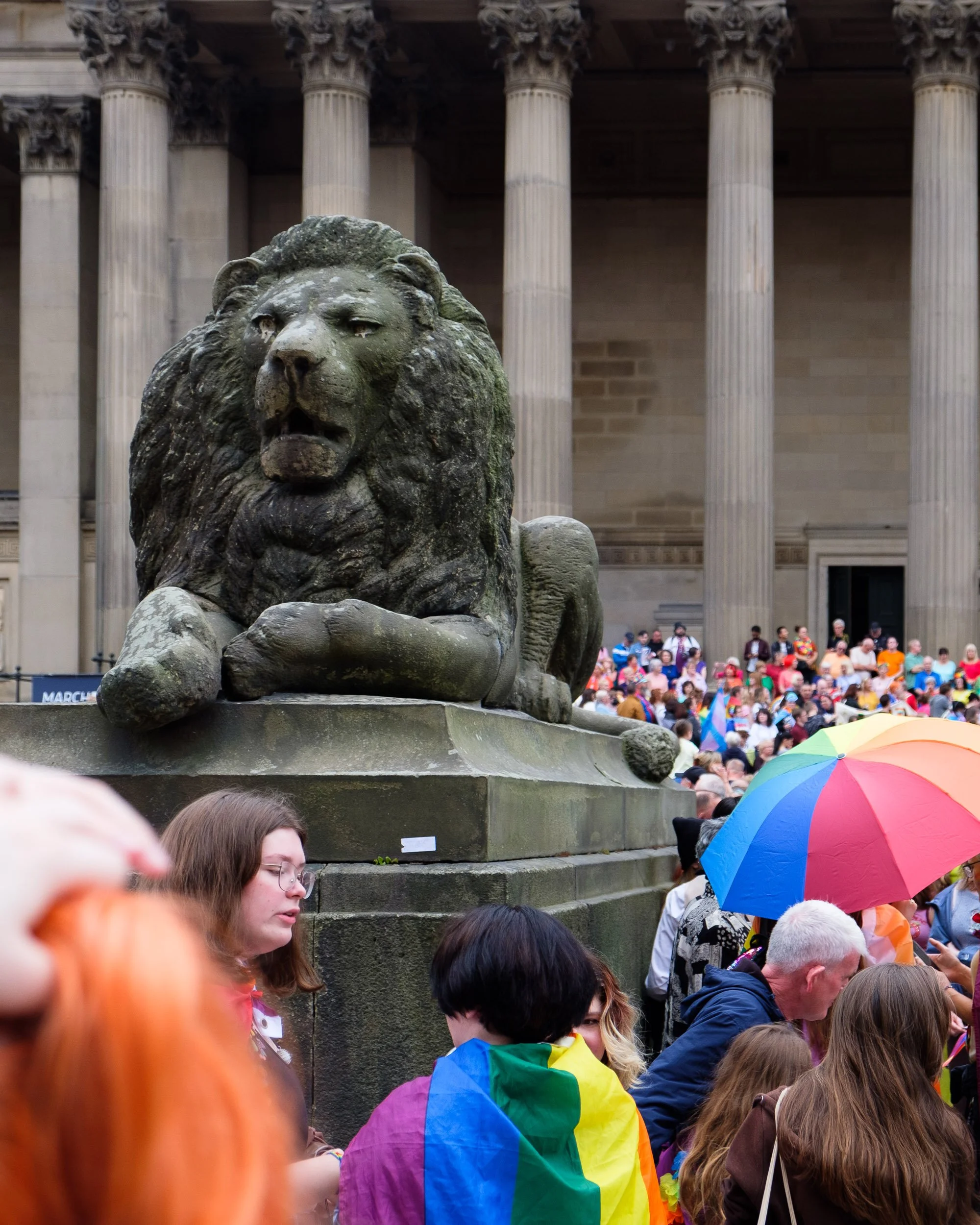 Bronze lion statue in front of a crowd of people holding rainbow flags and umbrellas, with large columns of a building in the background.