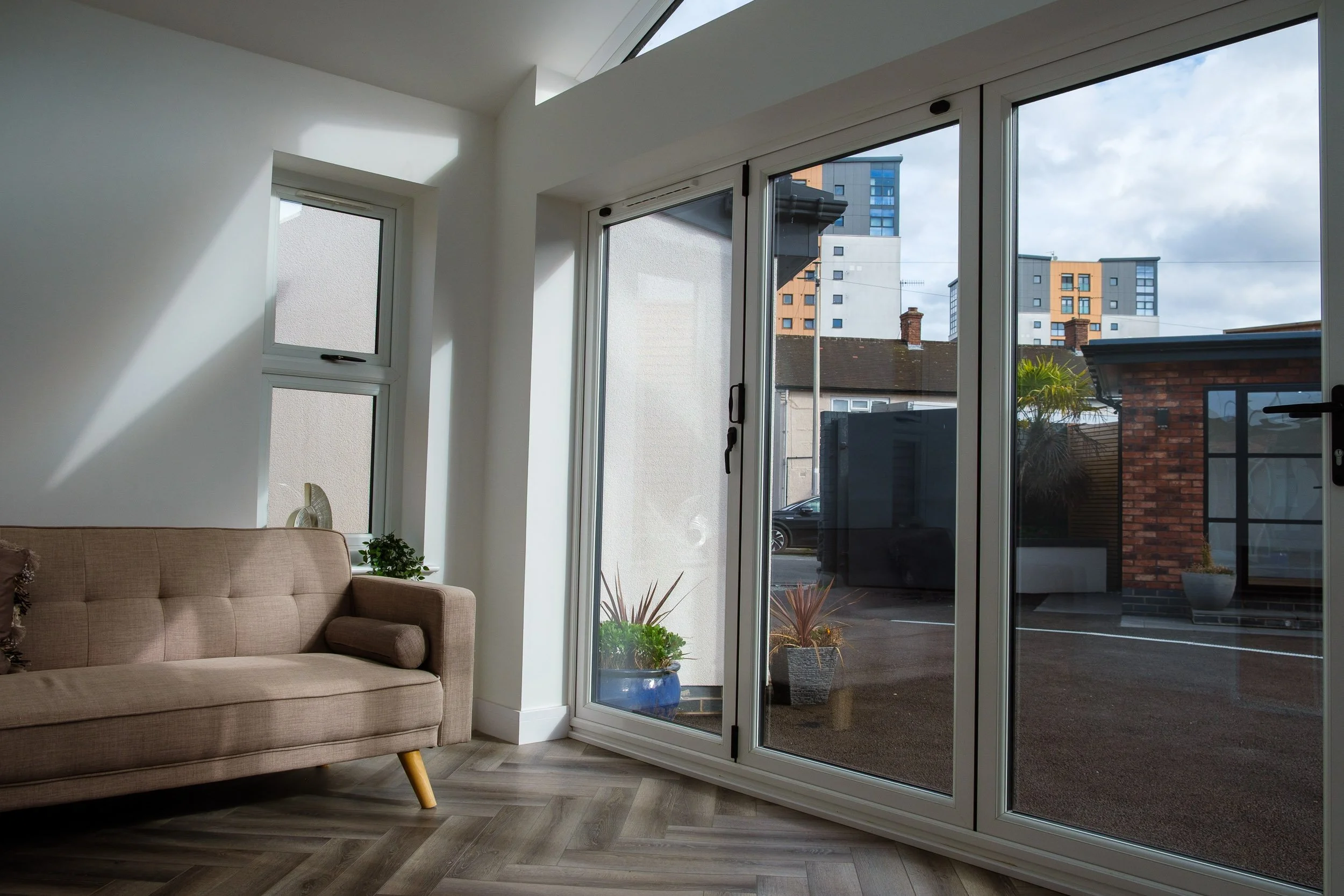 Living room with a beige sofa, a small plant, and large floor-to-ceiling glass sliding doors leading outside to a parking lot and city buildings.