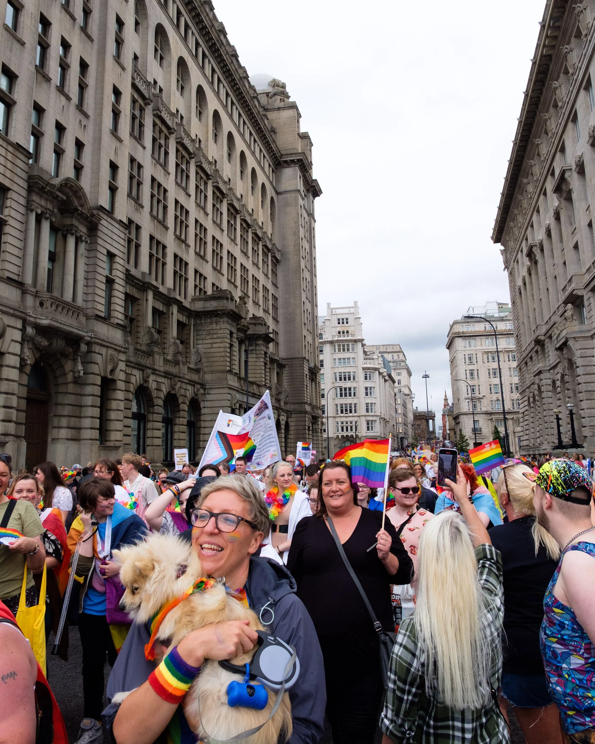 Crowd of people celebrating at a Pride parade, holding rainbow flags and wearing rainbow accessories on a city street with tall buildings.
