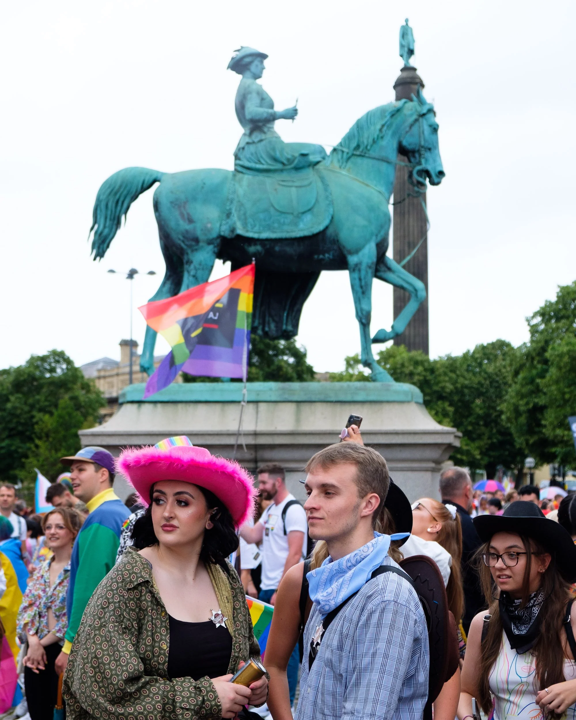 People gathered at a pride parade in front of a large equestrian statue, some holding rainbow flags and wearing colorful clothing and accessories.