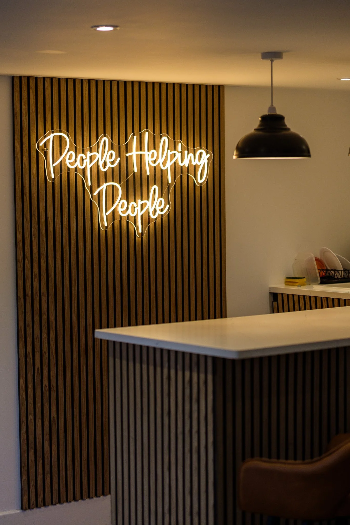 Neon sign on a wooden slatted wall that reads 'People Helping People' with a heart shape outline. Part of a kitchen counter with a brown chair in the foreground.