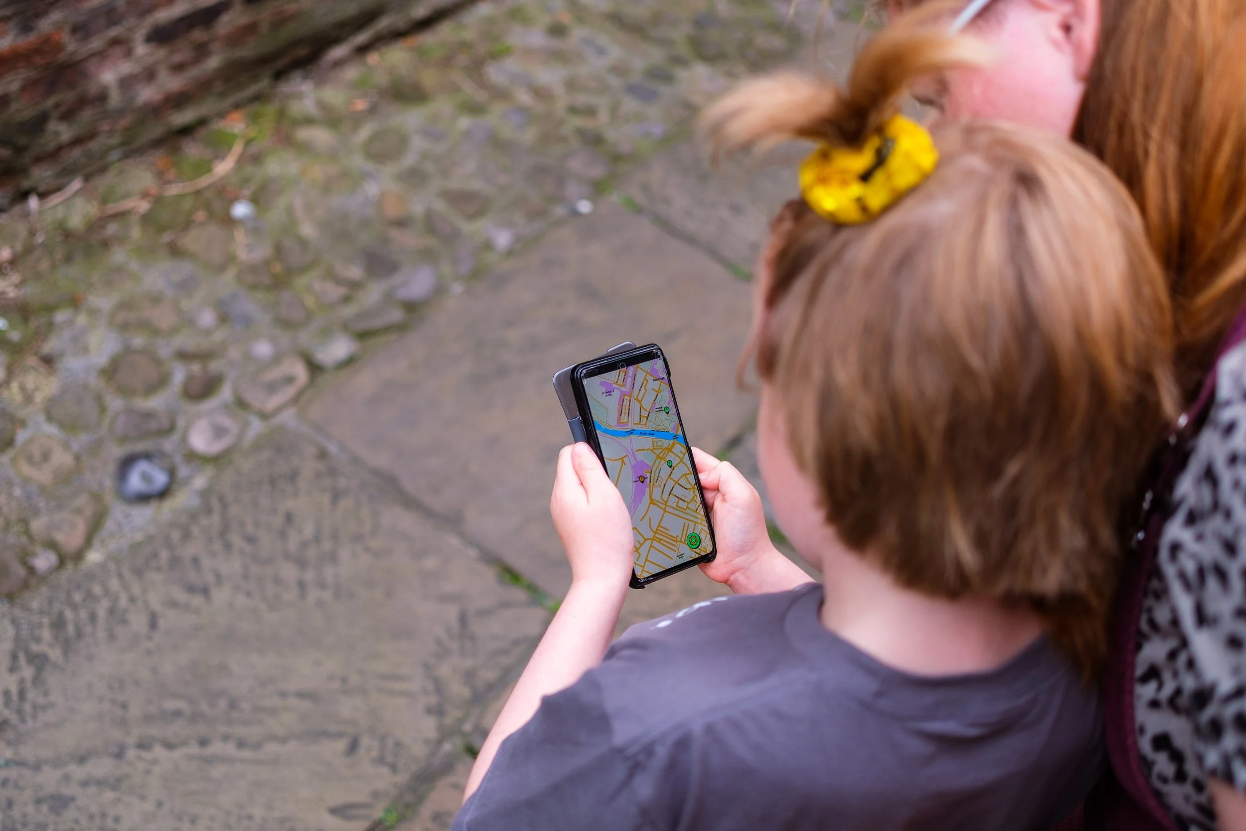 A person with short red hair holding a smartphone with a map app open, standing on a stone pathway next to a woman with long red hair.