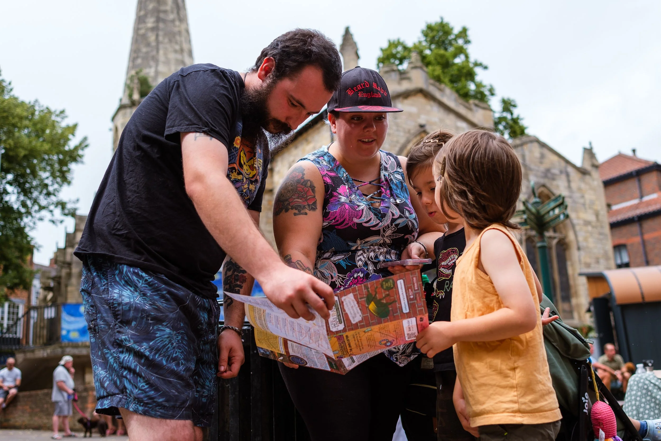 Four people, two adults and two children, are gathered around a map or brochure outside in front of a historic stone building with turrets, trees, and other people in the background.