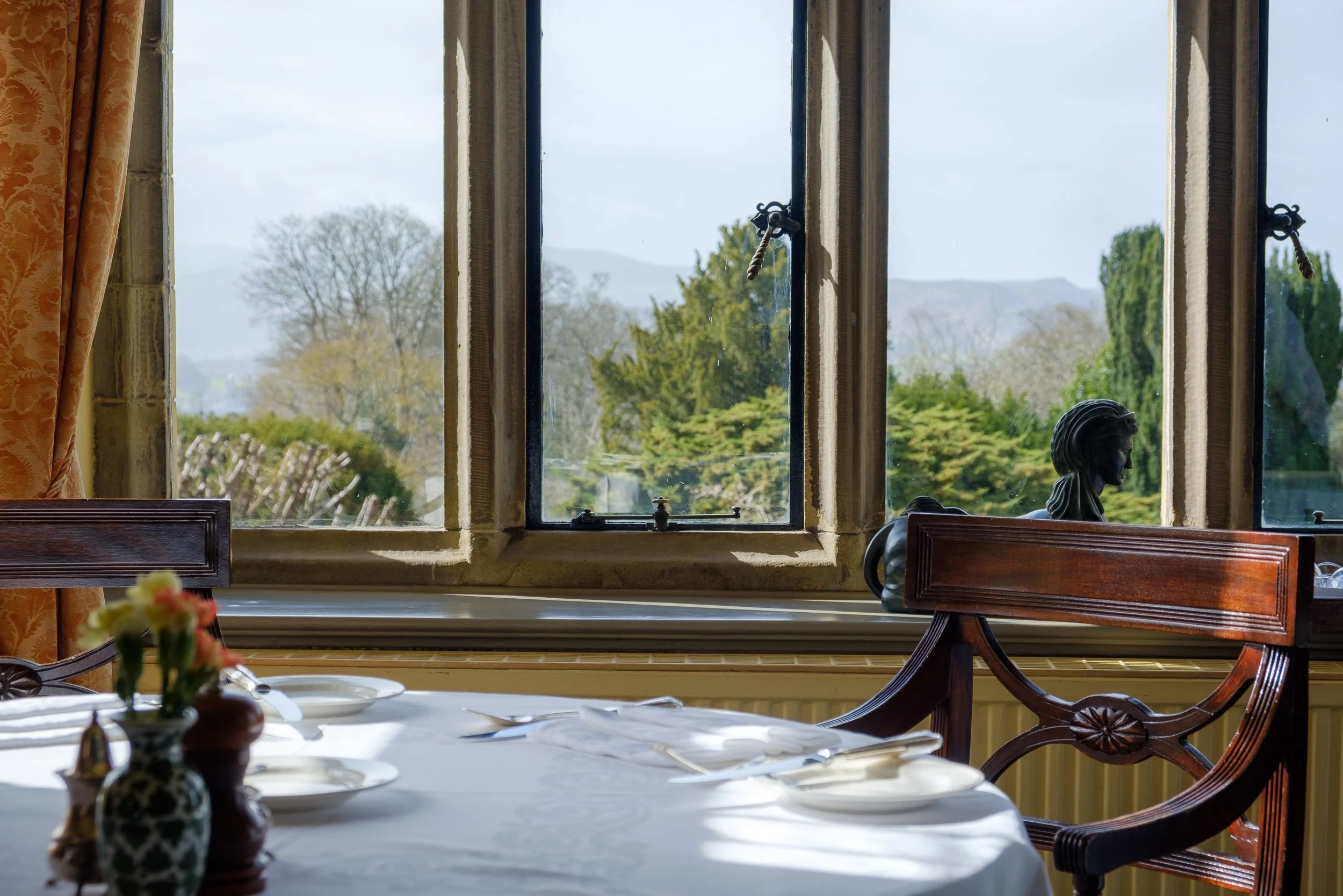 Interior view of a dining room with a table set with white plates, silverware, and small flower vases, overlooking a landscape through an open window showing trees and hills.