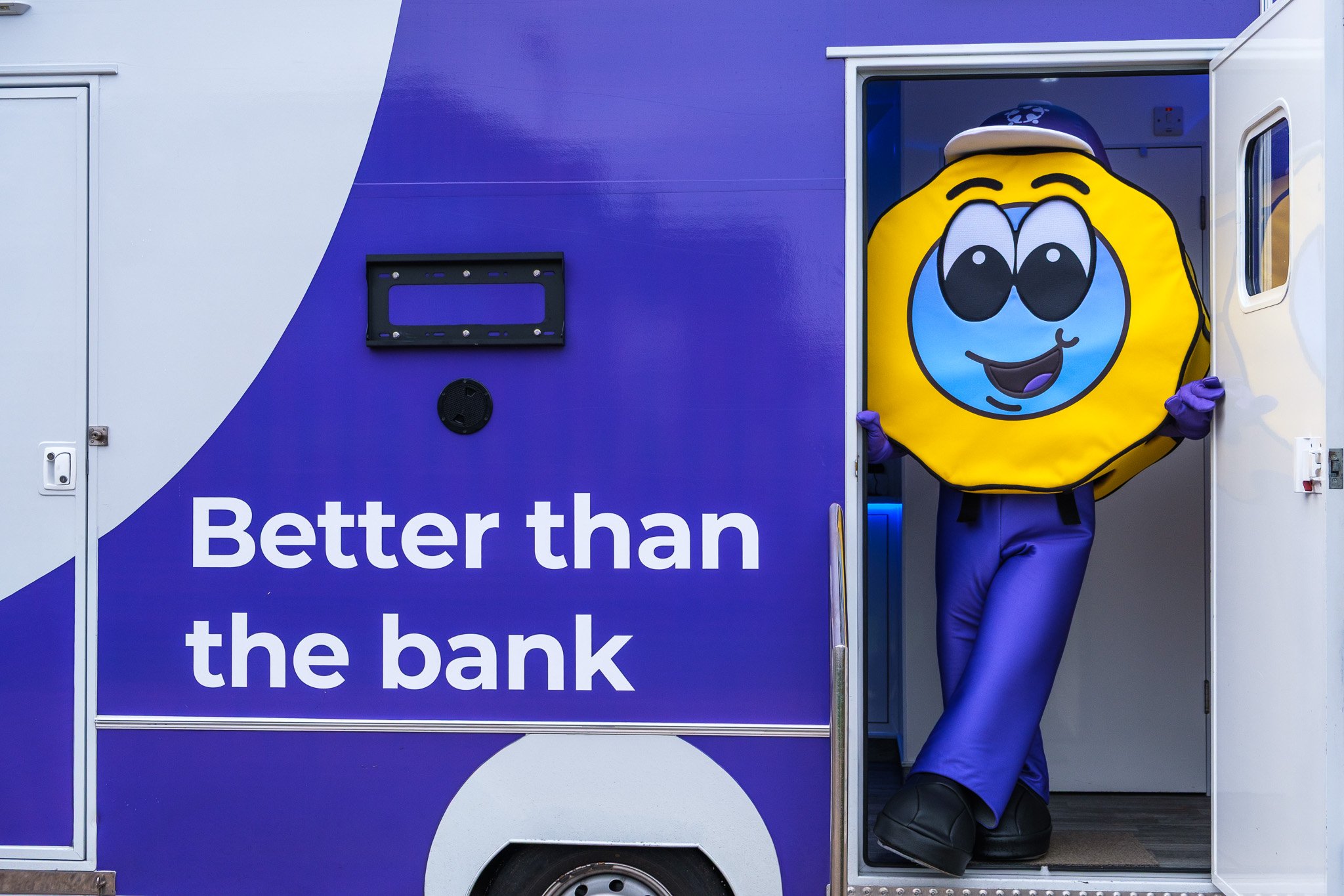A person in a yellow smiley face costume, wearing blue pants and a hat, is stepping out of a blue and white bank truck with the words "Better than the bank" written on it.
