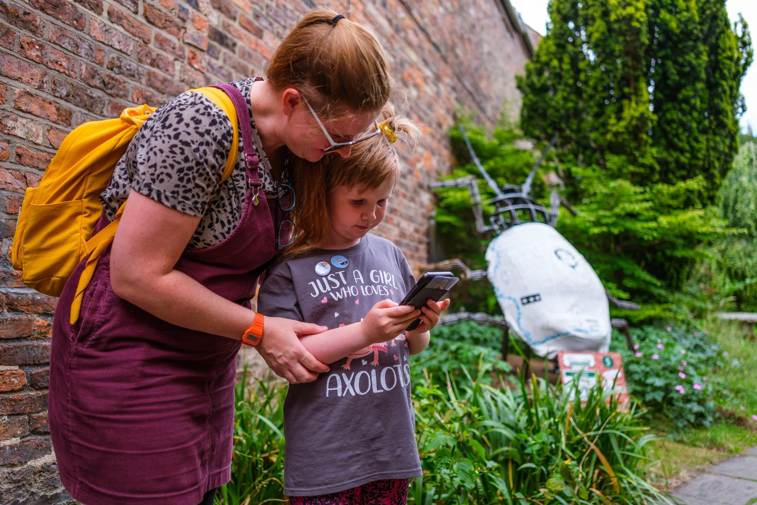 A woman and girl standing outside near a brick wall, looking at a smartphone together. The girl is wearing a gray shirt that says 'Just a girl who loves axolotls' and has buttons on her shirt. In the background, there's a white robot sculpture with b