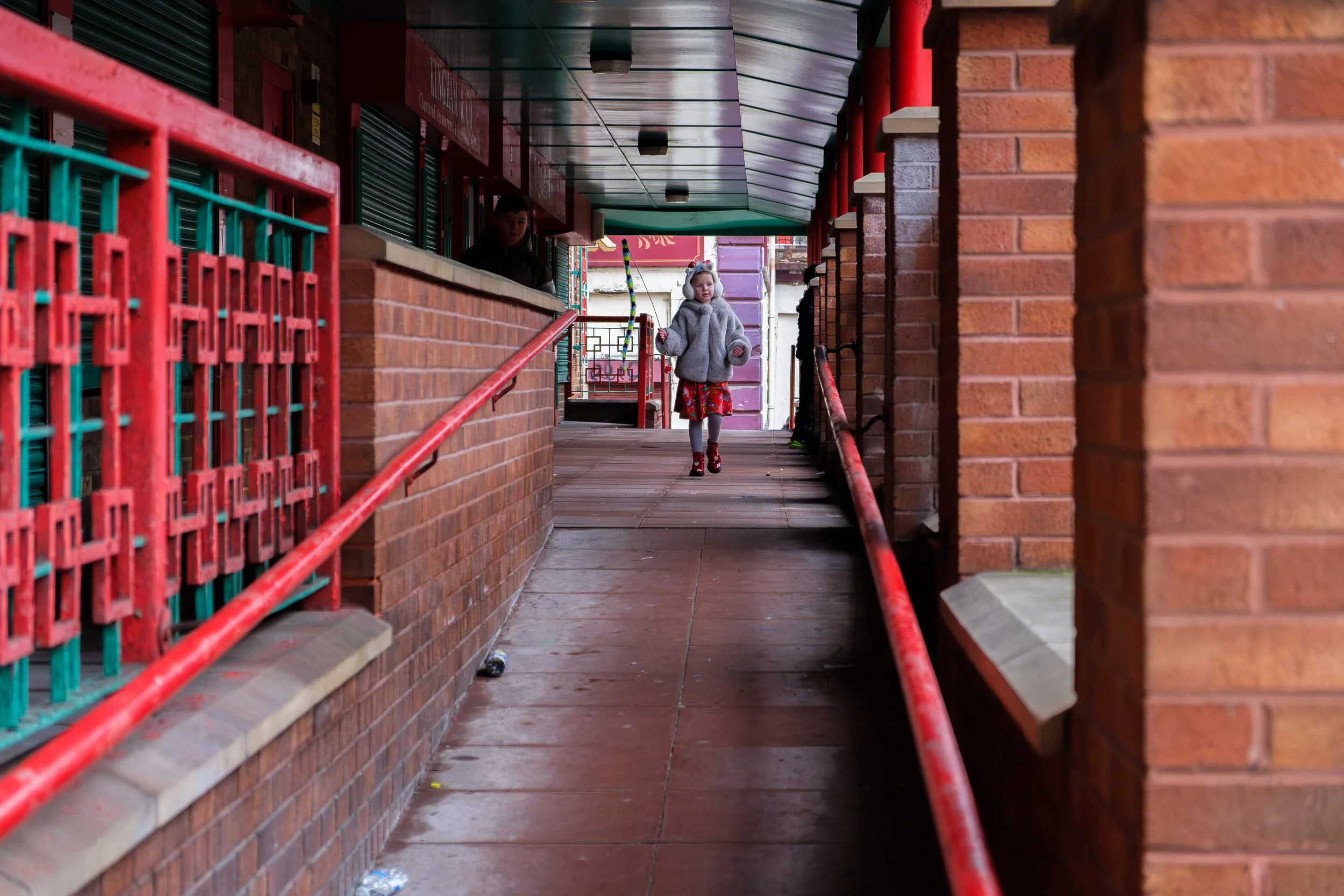 Young girl walking through a narrow outdoor alleyway between brick buildings with red handrails, wearing a large gray jacket and red patterned shoes.