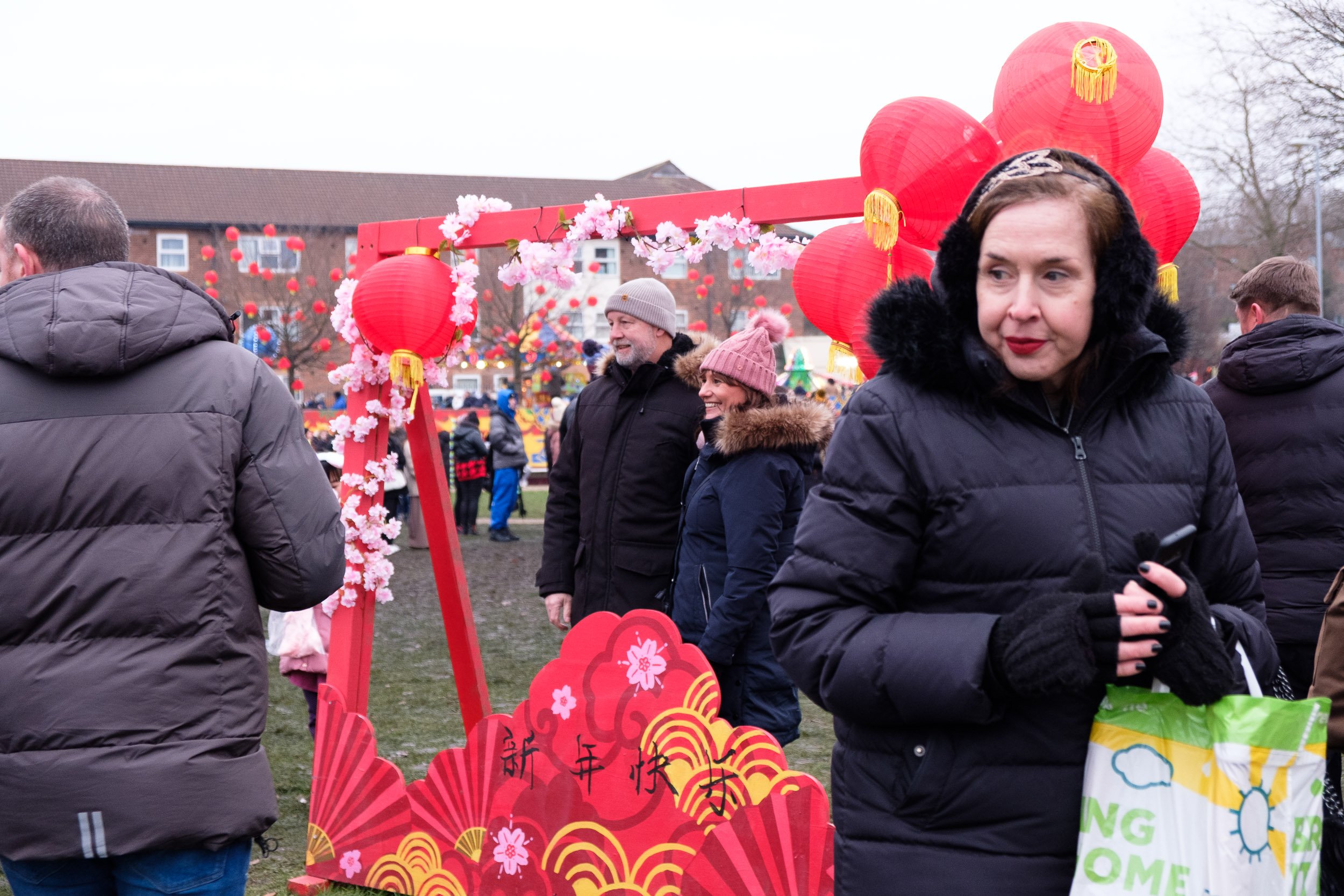 People gathered around a red decorative arch with red lanterns and pink flowers at an outdoor celebration, likely for Chinese New Year.