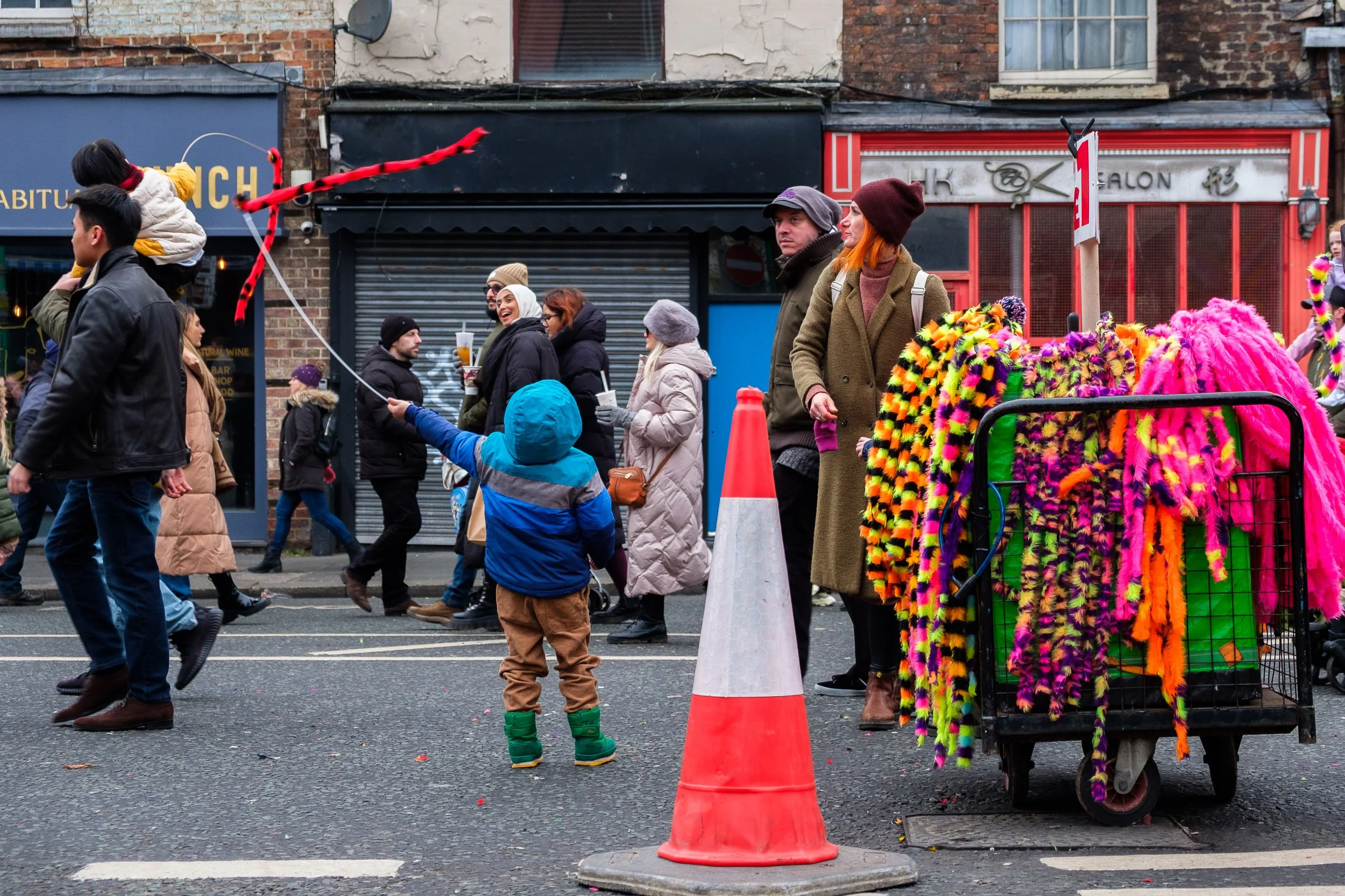 People walking on a city street during a festive event, with a vendor selling colorful feather boas, and some individuals holding drinks, while a young child in a blue jacket and green boots reaches out to a balloon.