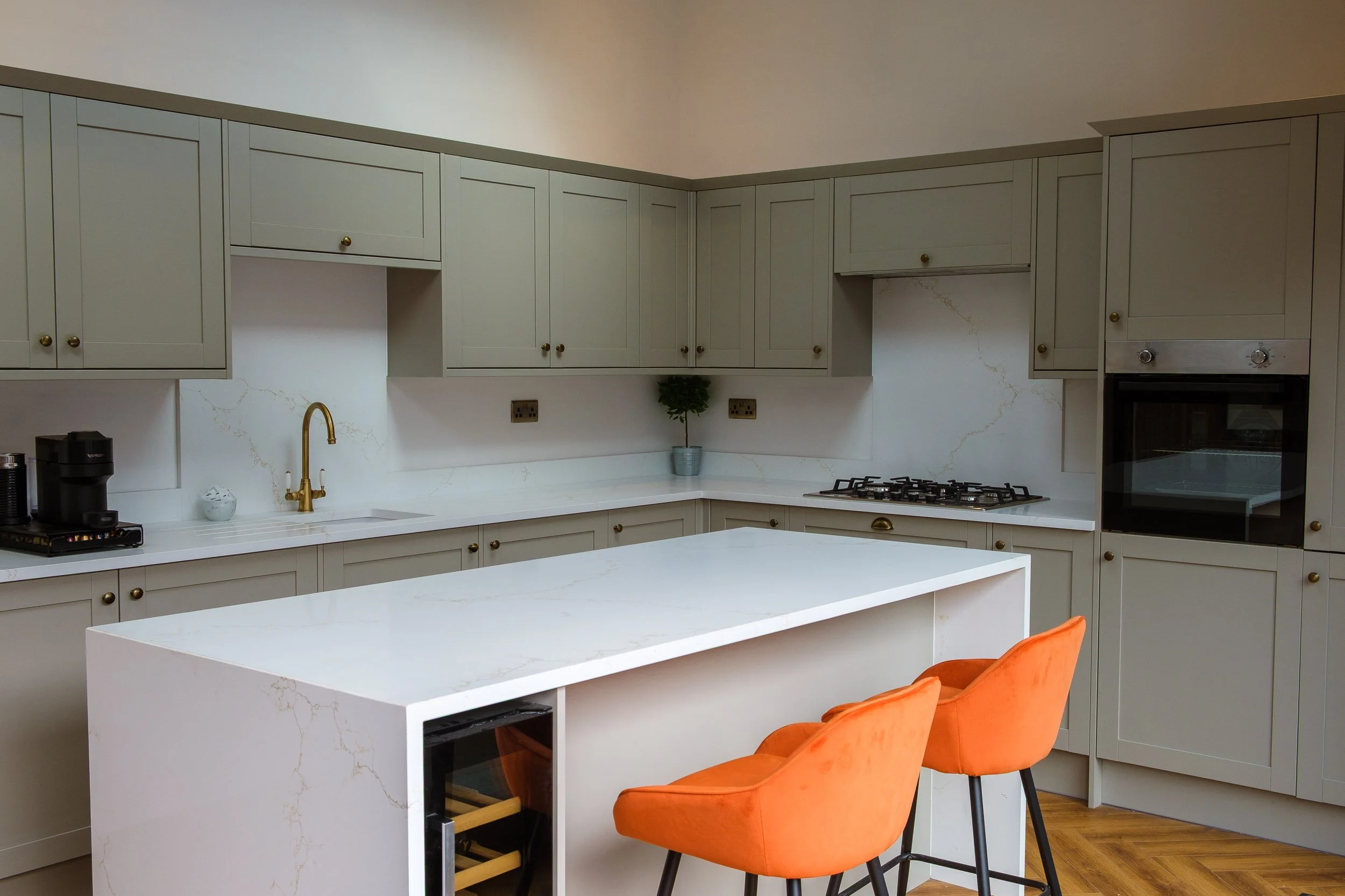 Modern kitchen with white marble countertops, gray cabinets, and an island. Orange bar stools are in front of the island, kitchen features a sink, stove, and built-in oven.