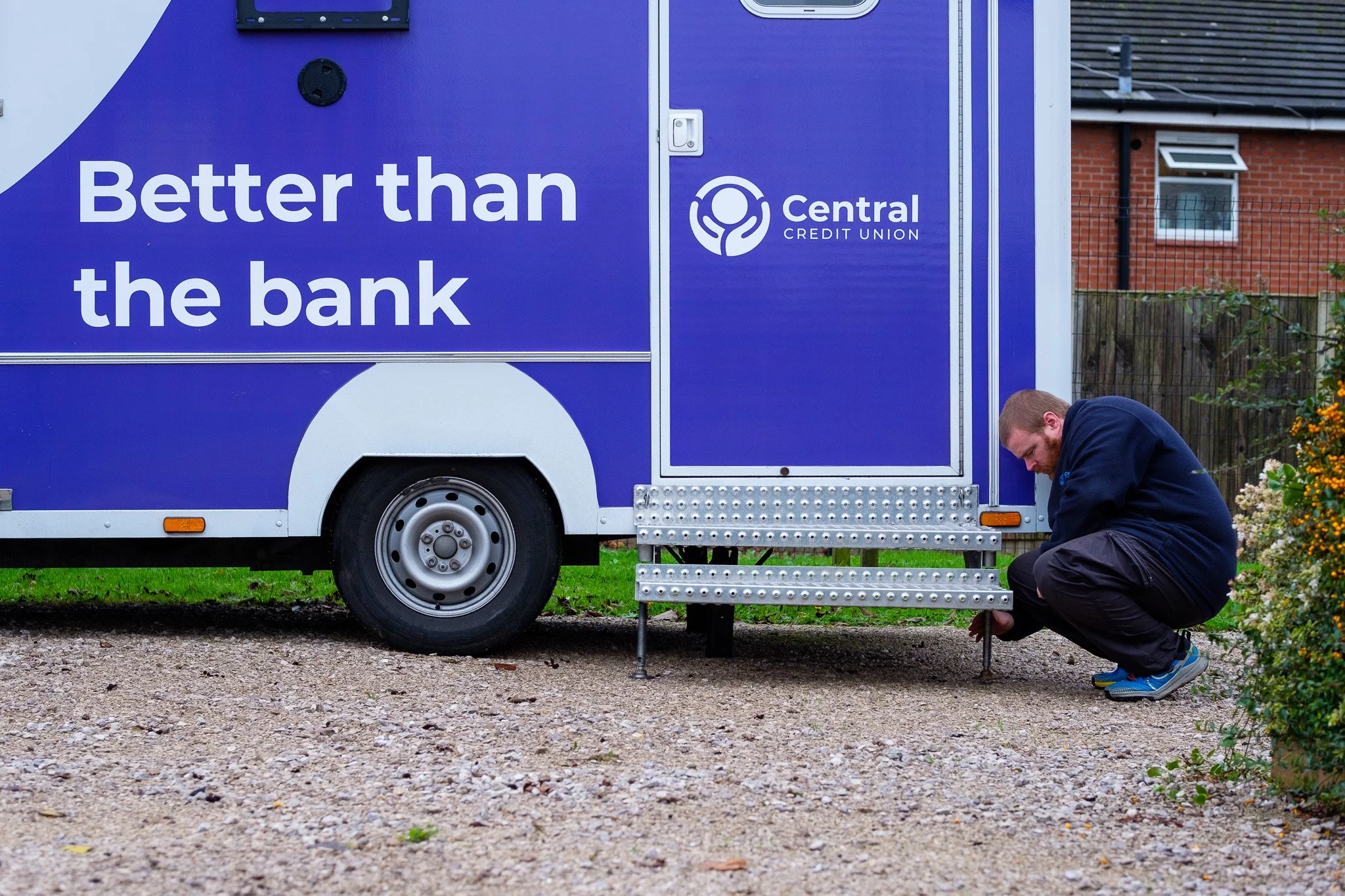 A man crouching next to a mobile bank van labeled 'Better than the bank' with the logo of Central Credit Union, adjusting or inspecting something near the vehicle's steps.