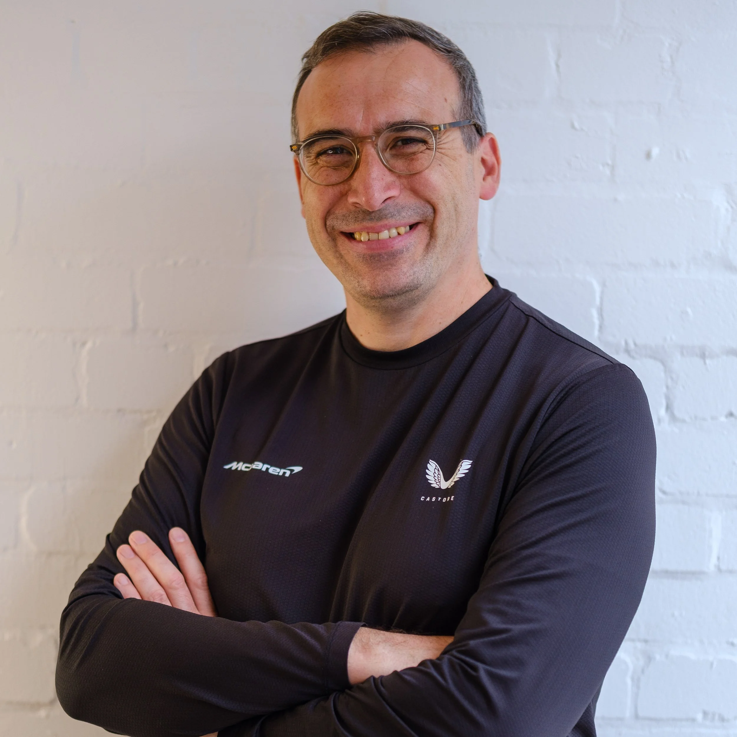 A man wearing glasses and a black sports shirt with logos, standing with arms crossed in front of a white brick wall.