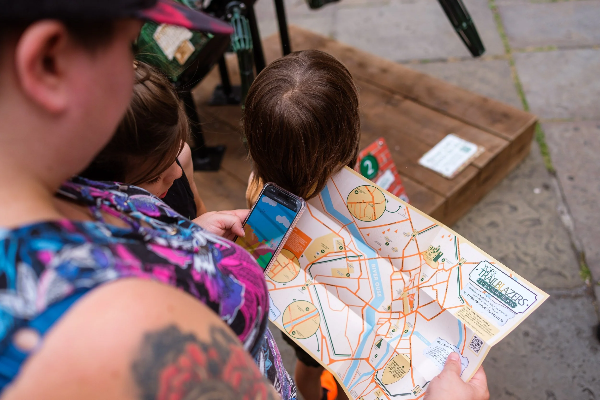 Two women and a child looking at a city map of York, with one woman holding a phone. The scene is outdoors, on a sidewalk near a wooden platform.