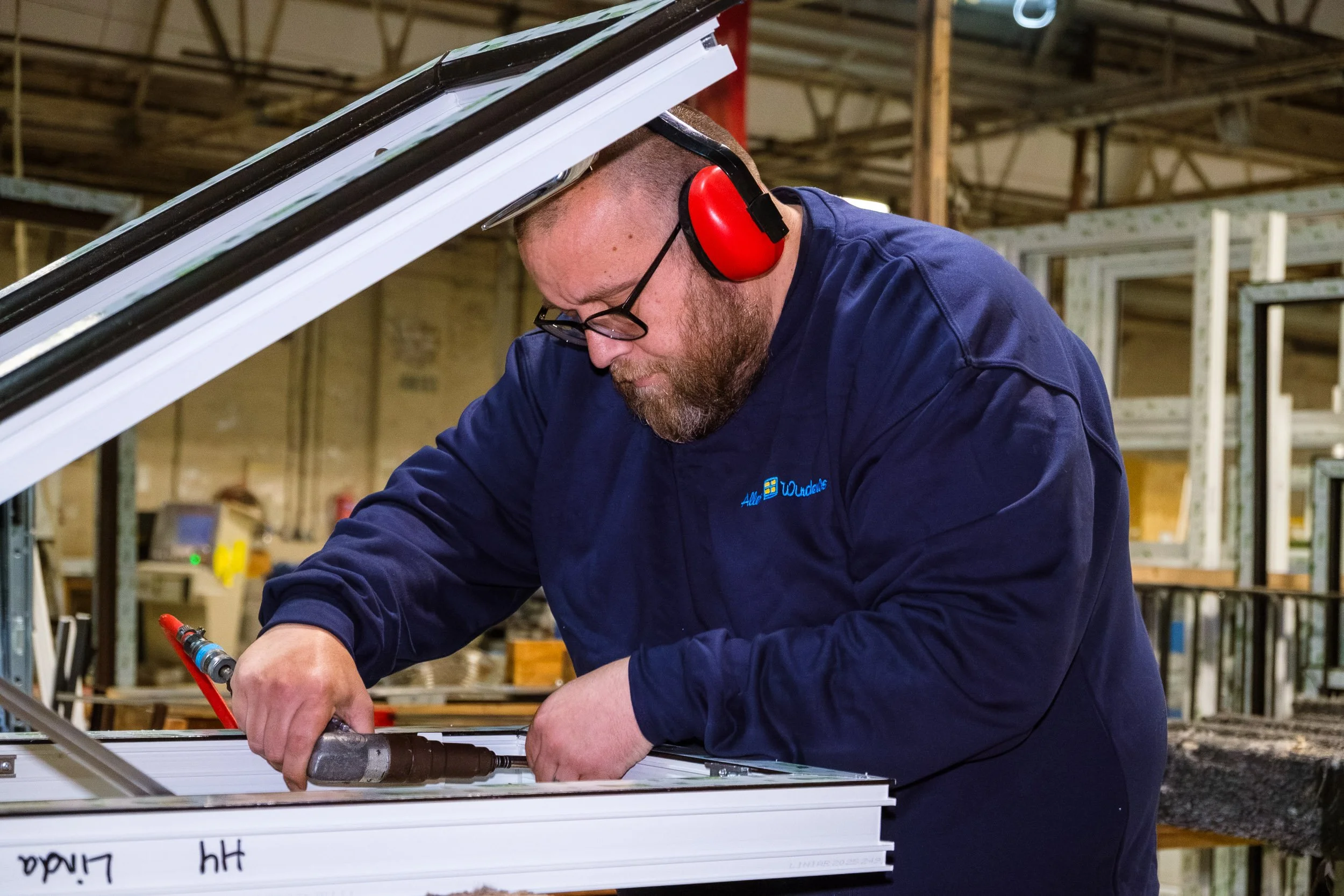 A man working on a construction or manufacturing project, wearing red ear protection and glasses, using a power tool on a white metal frame.