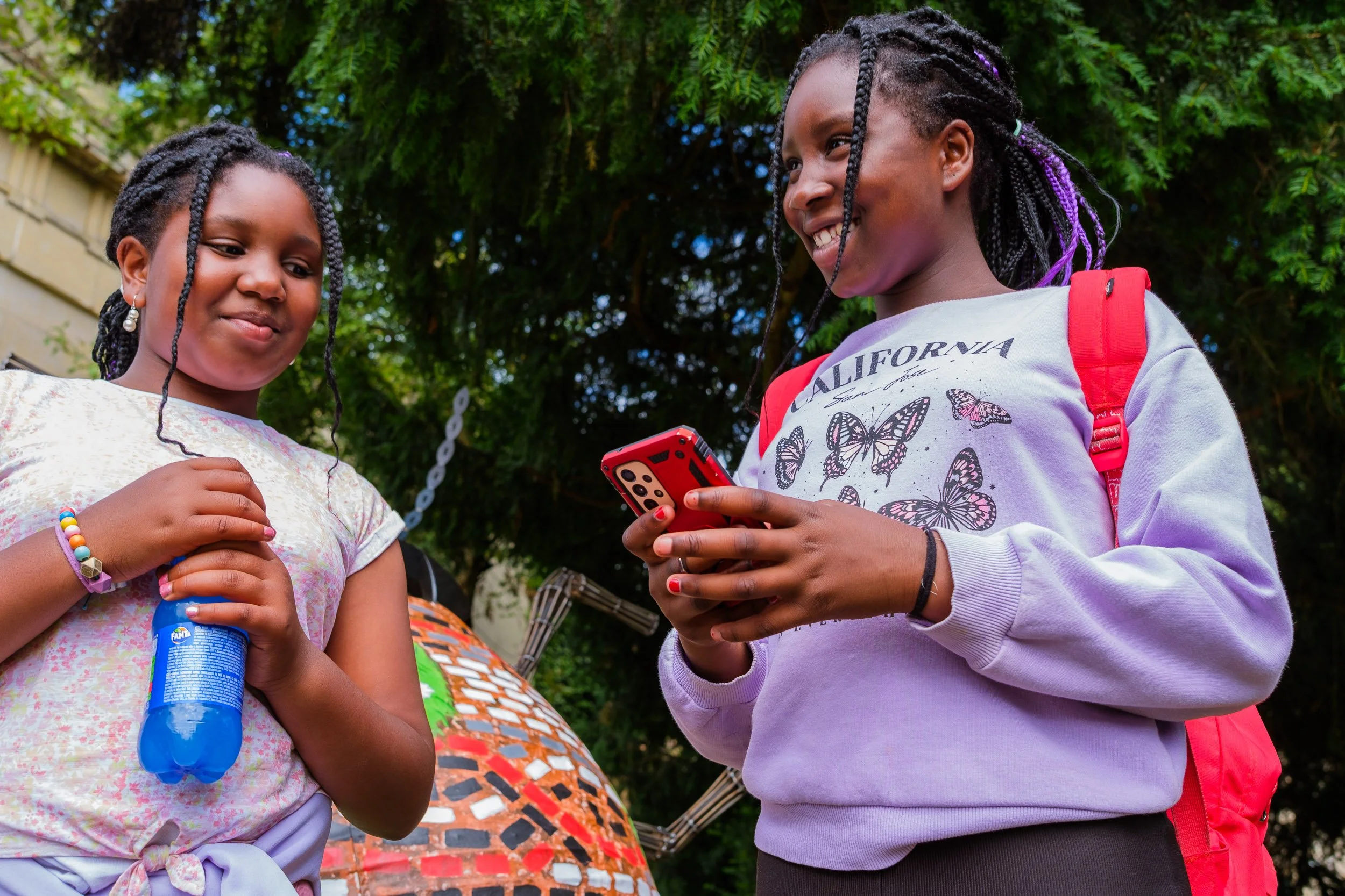 Two young girls with braided hair and backpacks outdoors, one holding a phone with a butterfly display case, smiling and looking at the screen, surrounded by green trees.