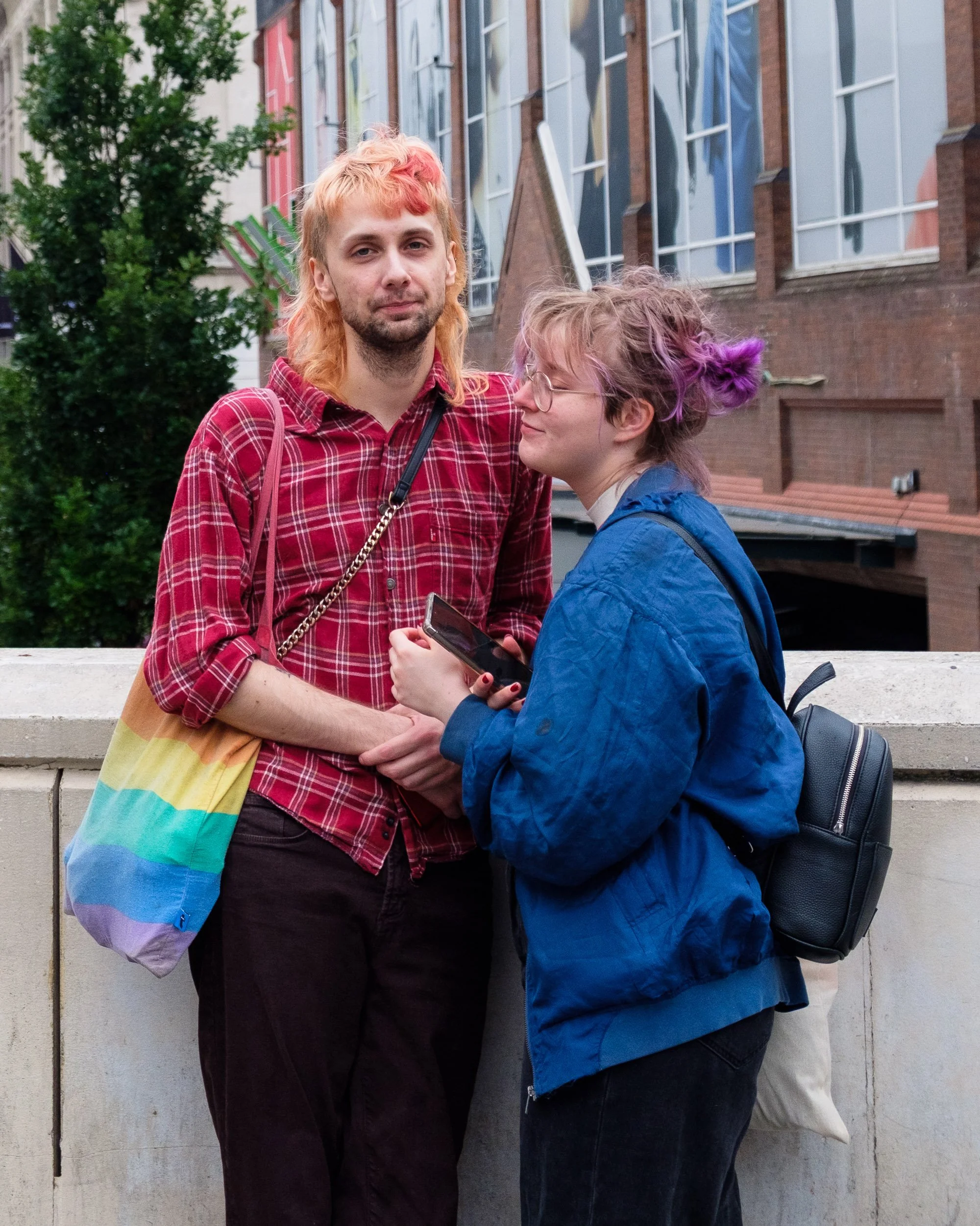 Two young people with dyed hair standing on a city street, one wearing a red plaid shirt and the other a blue jacket, with backpacks, engaged in conversation.