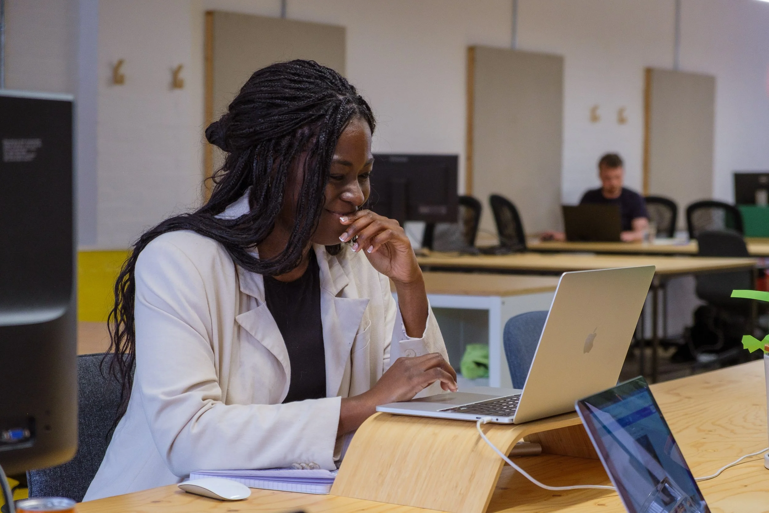 Woman with braided hair working on a silver laptop in a modern office, with a notebook and a mouse on the desk, and a man working on a computer in the background.
