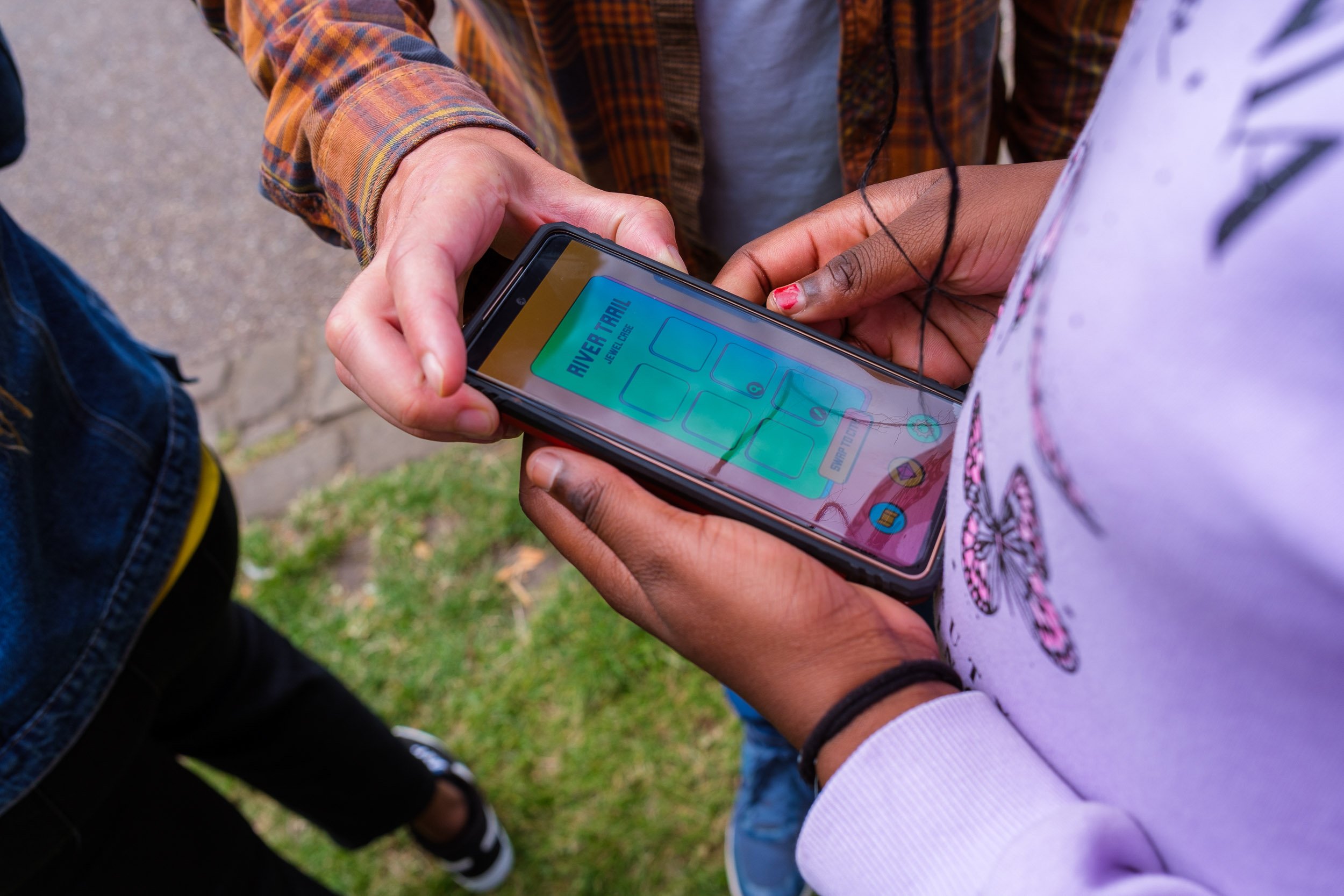 A person holding a smartphone displaying a trail app with icons for different locations and options, outdoors on grass and pavement.