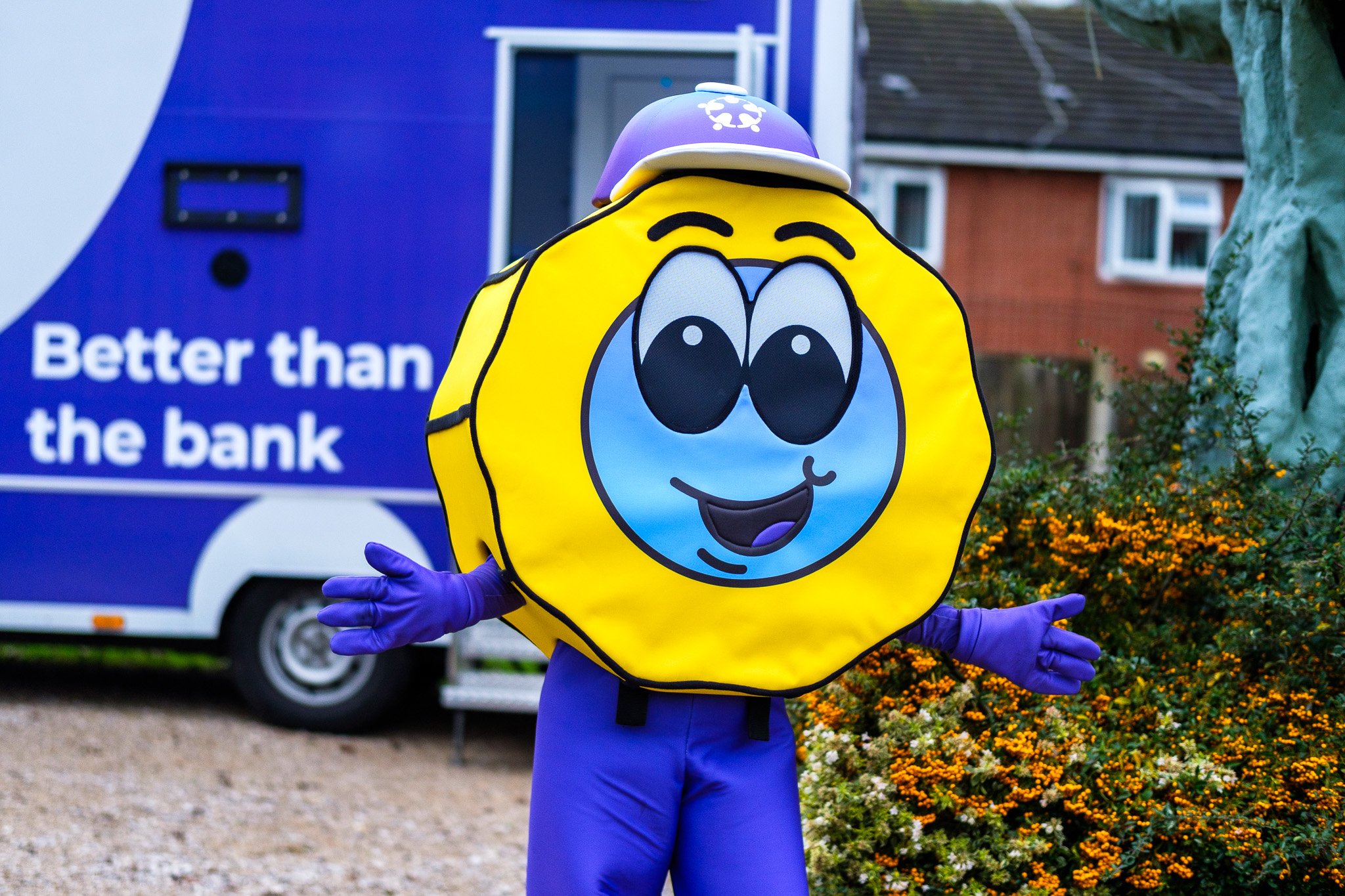 A person dressed in a lemon mascot costume with a bright yellow body, large cartoon eyes, a smiling face, purple gloves, and a purple helmet. The mascot is standing outdoors in front of a blue bank truck with the sign 'Better than the bank.' in the b