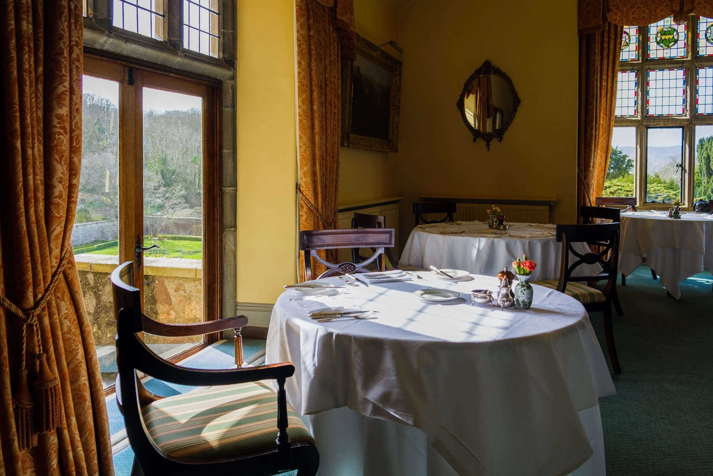 A cozy dining room with round tables covered in white tablecloths, set with plates and cutlery, and decorated with flowers. Large windows with stained glass and orange curtains let in natural light and offer a view of a garden outside.