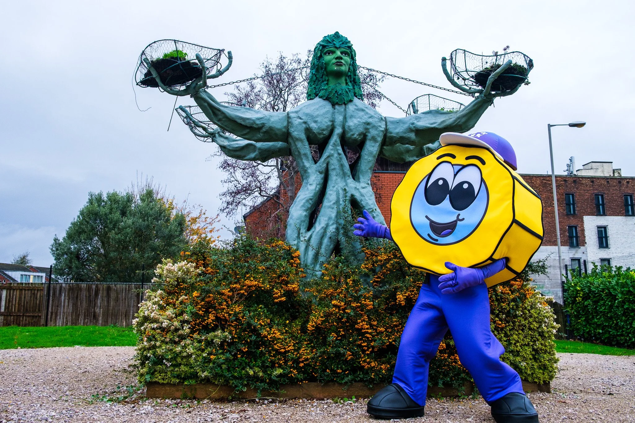 A person in a mascot costume resembling a yellow coin with a smiling face, arms, and purple gloves standing in front of a large sculpture of a woman with multiple arms holding baskets, with a cloudy sky above, bushes, a fence, and buildings in the ba