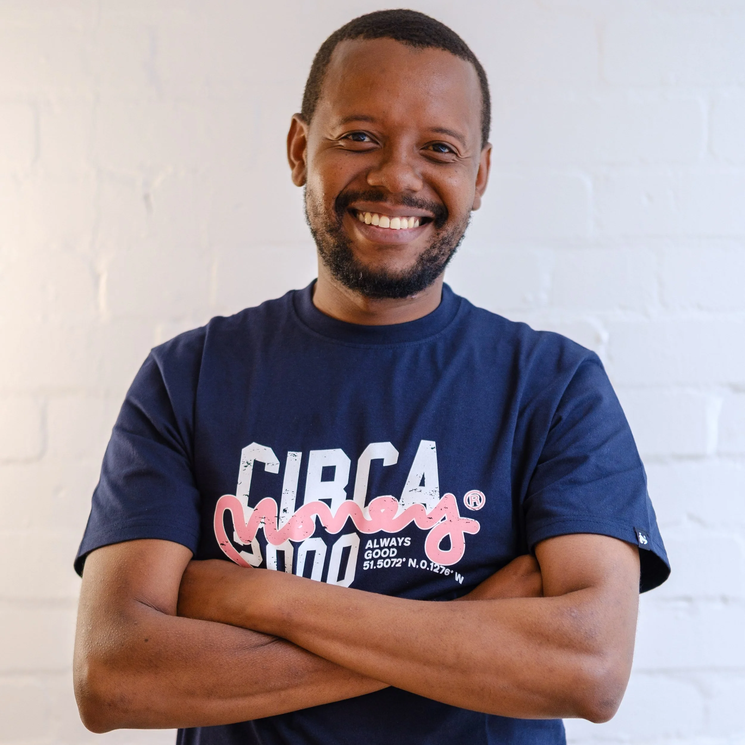 A smiling man with a beard and mustache standing with arms crossed against a white brick wall, wearing a navy blue T-shirt with pink and white text.