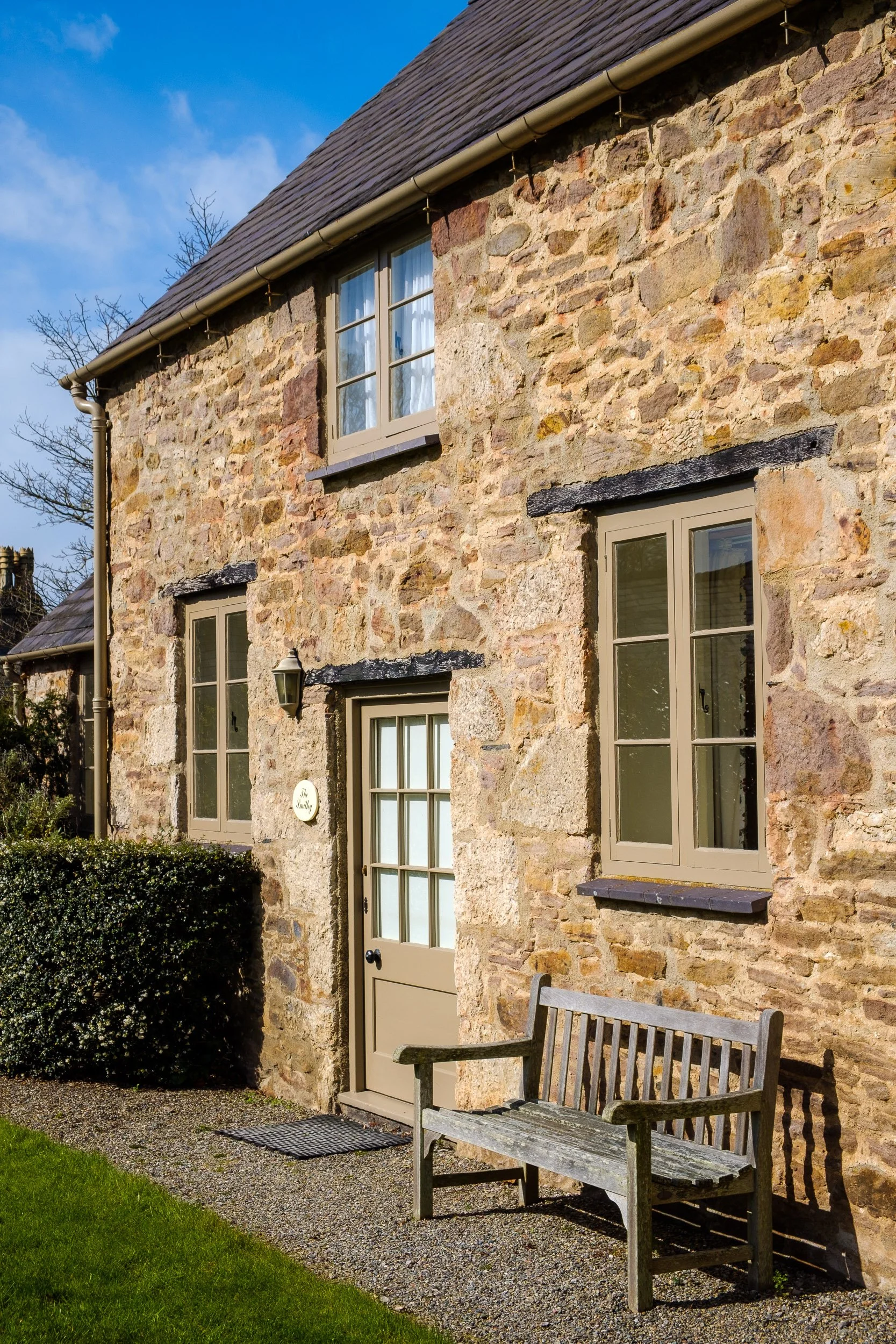 A stone house with beige window frames, a door with glass panes, a small outdoor bench, and a hedge, under a blue sky.