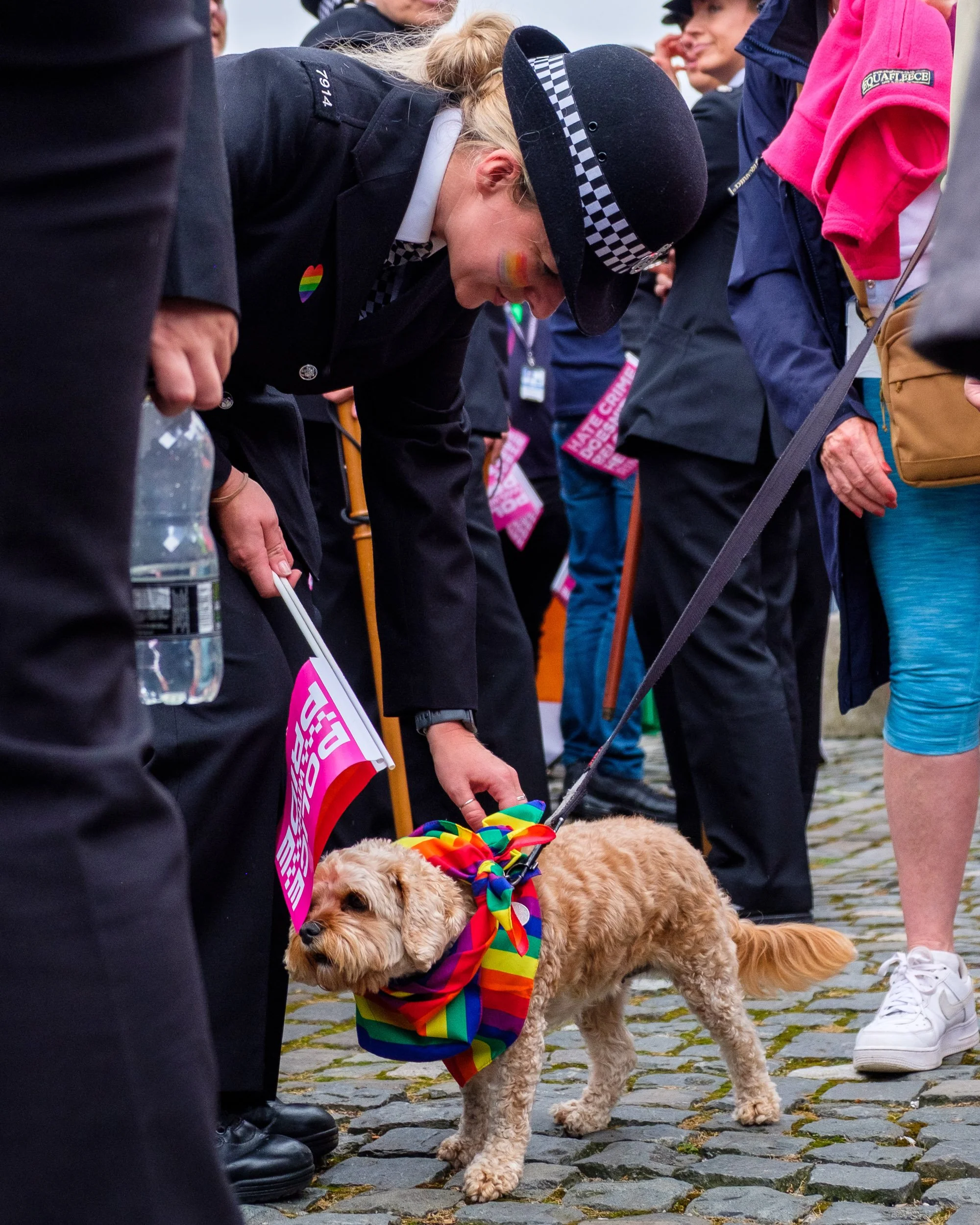 A woman in a police uniform bows down to a dog wearing a rainbow-colored bandana, surrounded by people at a pride event.