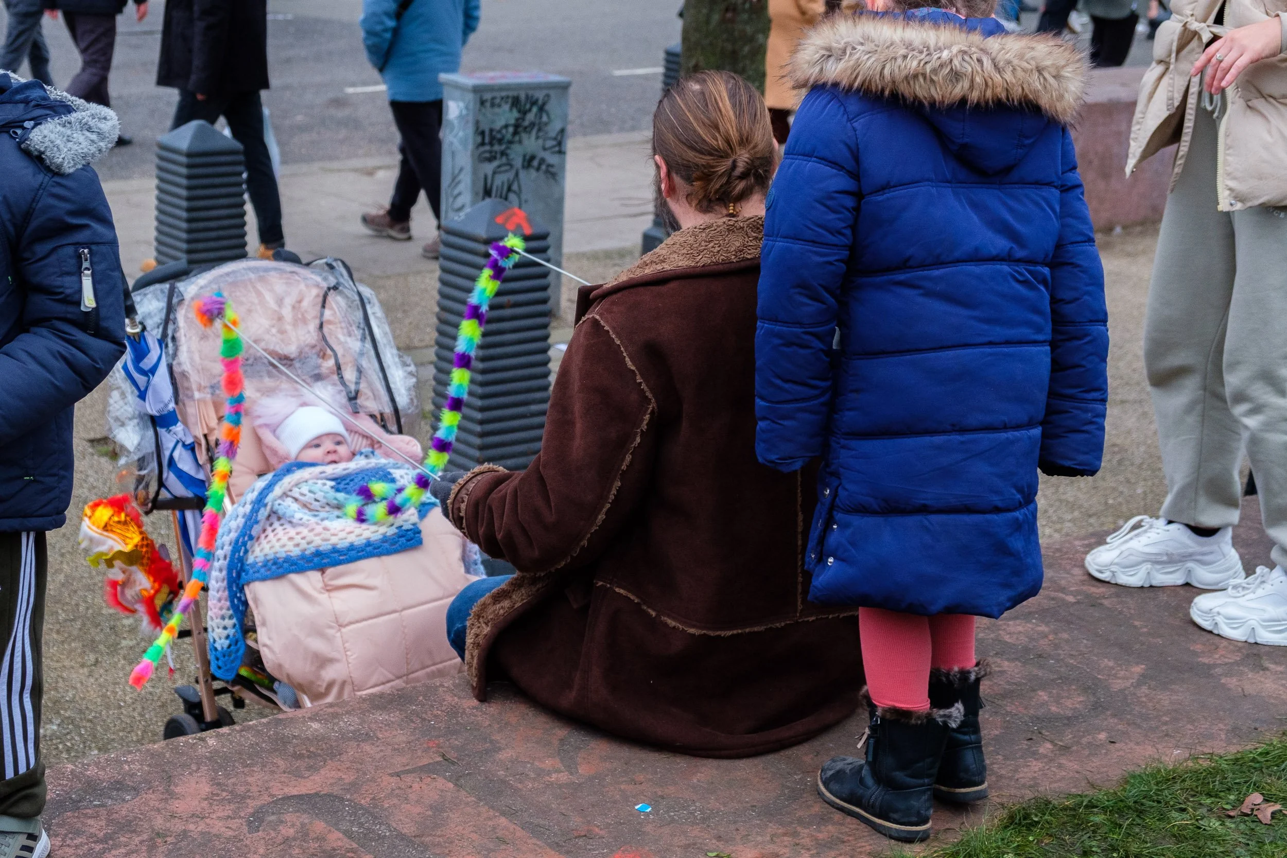A woman with brown hair tied back sitting on a ledge, holding a colorful string with a baby in a stroller nearby. The baby is wrapped in a blanket, wearing a white hat, and looking at the woman. Several children and adults are gathered around in a pa