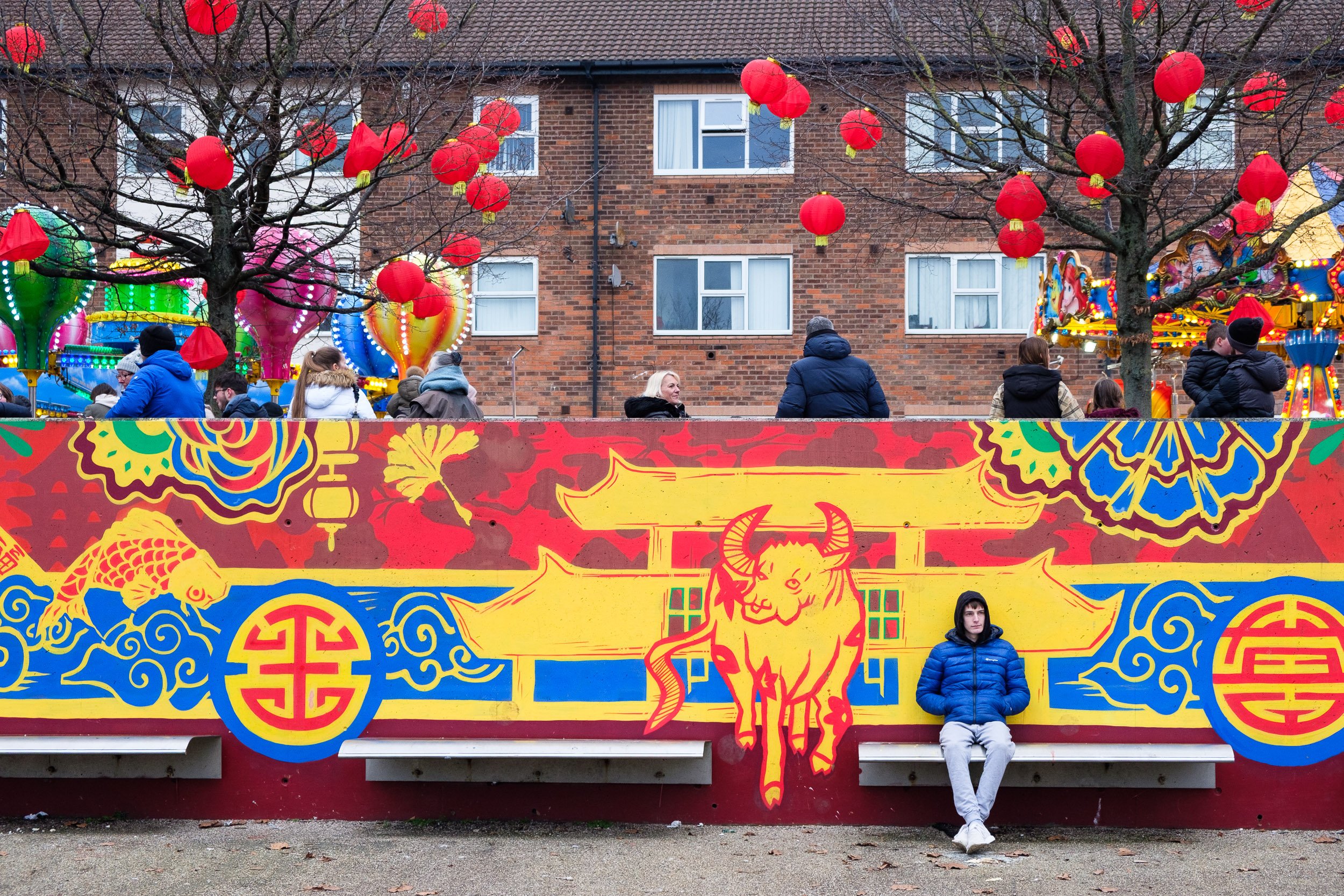 A colorful mural featuring a yellow bull with red details in front of a red background with blue and green patterns, with people at a carnival or fair behind a fence decorated with red lanterns hanging from leafless trees.