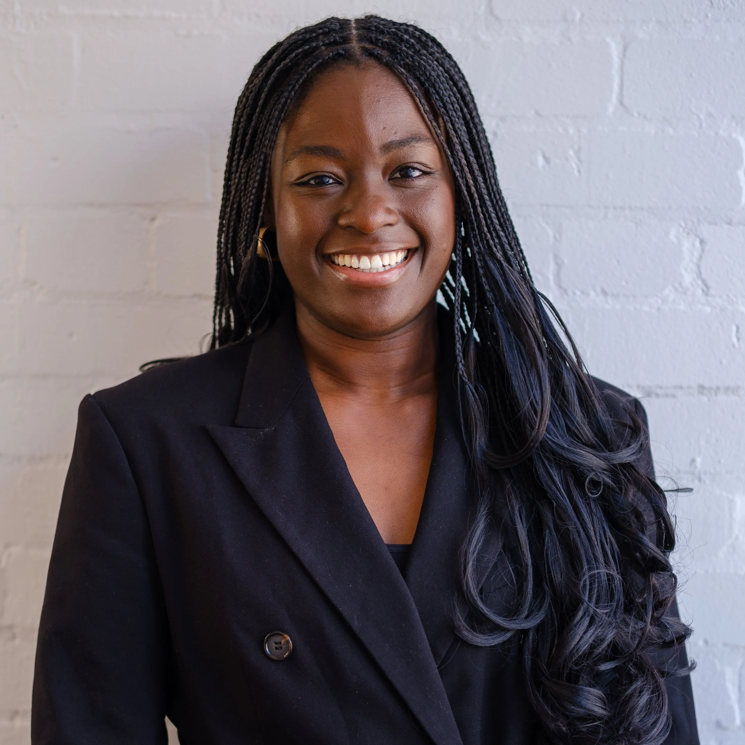 A portrait of a smiling Black woman with long braided hair, wearing a black blazer, standing in front of a white brick wall.