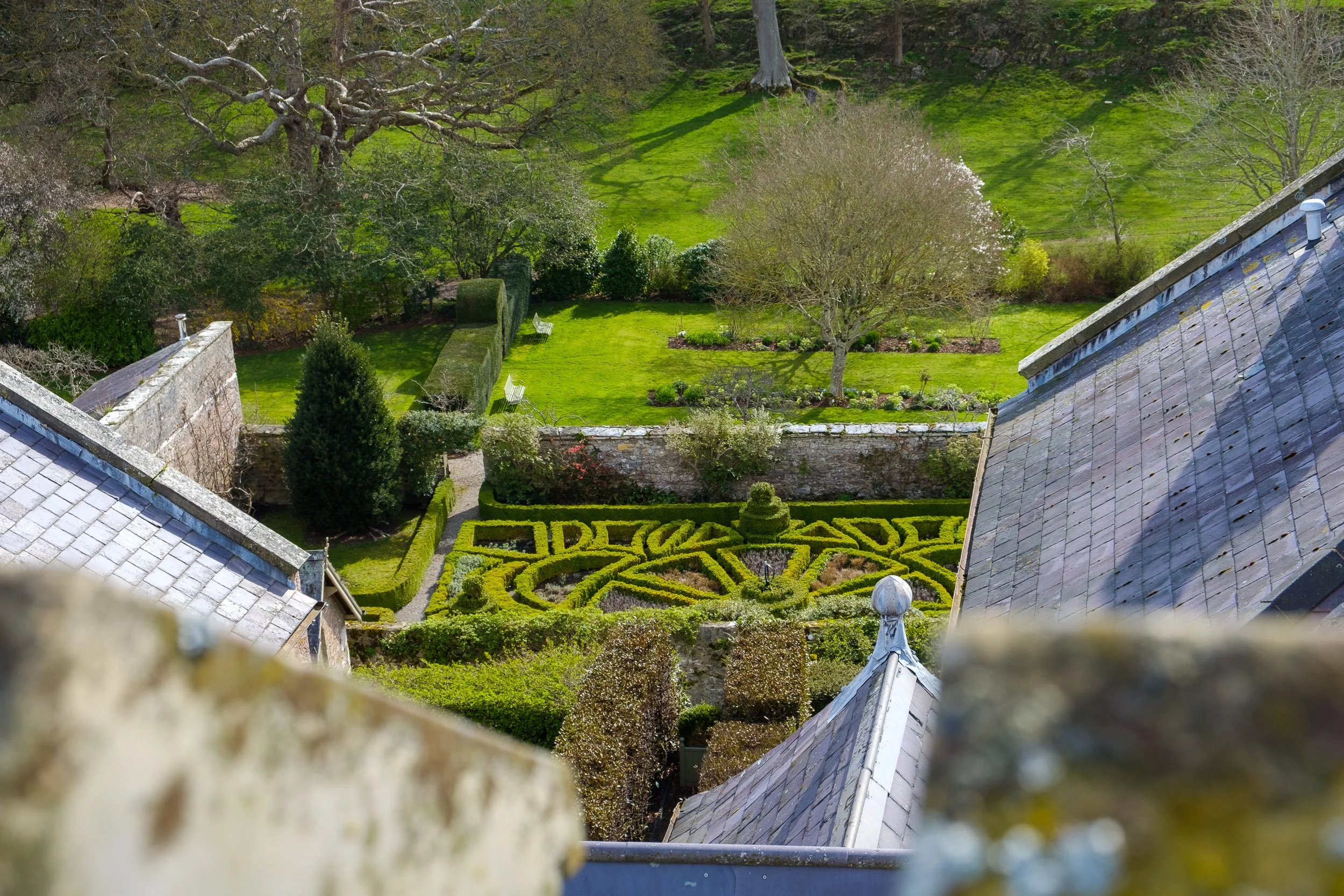A view of a well-maintained garden with trimmed hedges, a decorative maze, and a lawn surrounded by trees and stone walls, taken from above.