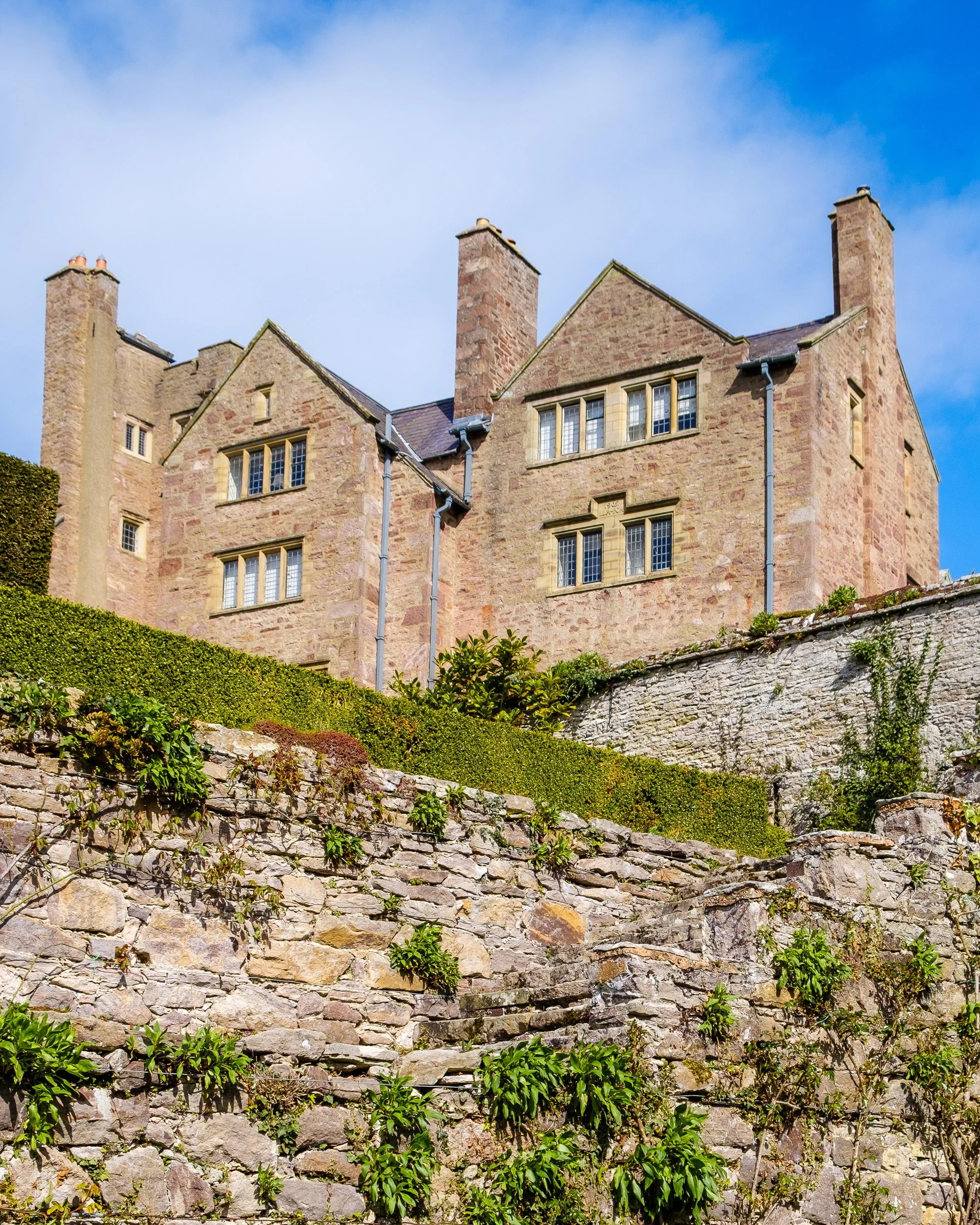 A historic stone castle with multiple chimneys and small windows, situated on a hill with stone and greenery, under a partly cloudy sky.
