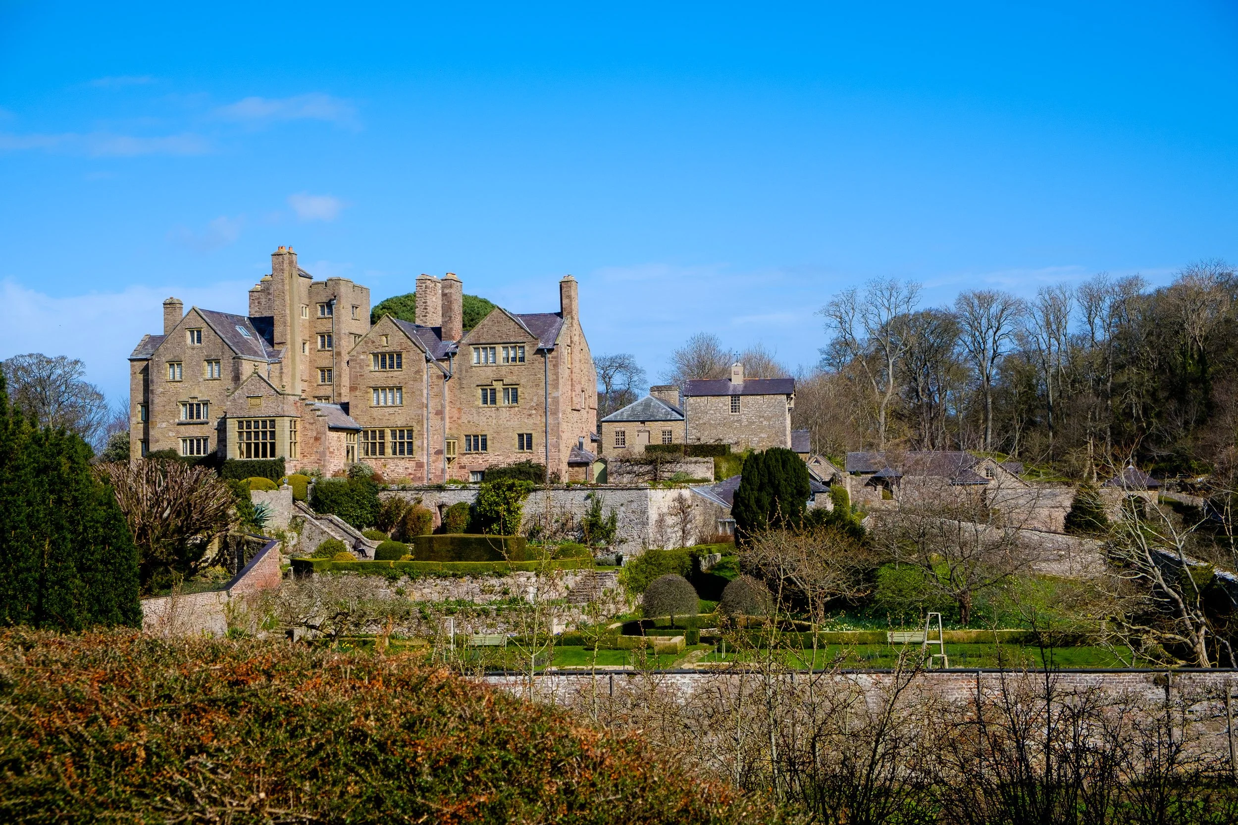 A large medieval-style stone castle on a hillside with surrounding gardens and trees under a blue sky.