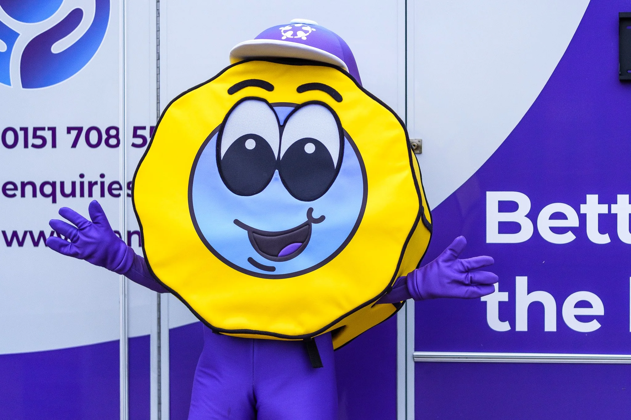 Person dressed in a lemon costume with cartoon lemon face, purple gloves, and a purple cap, standing in front of a purple and white billboard.