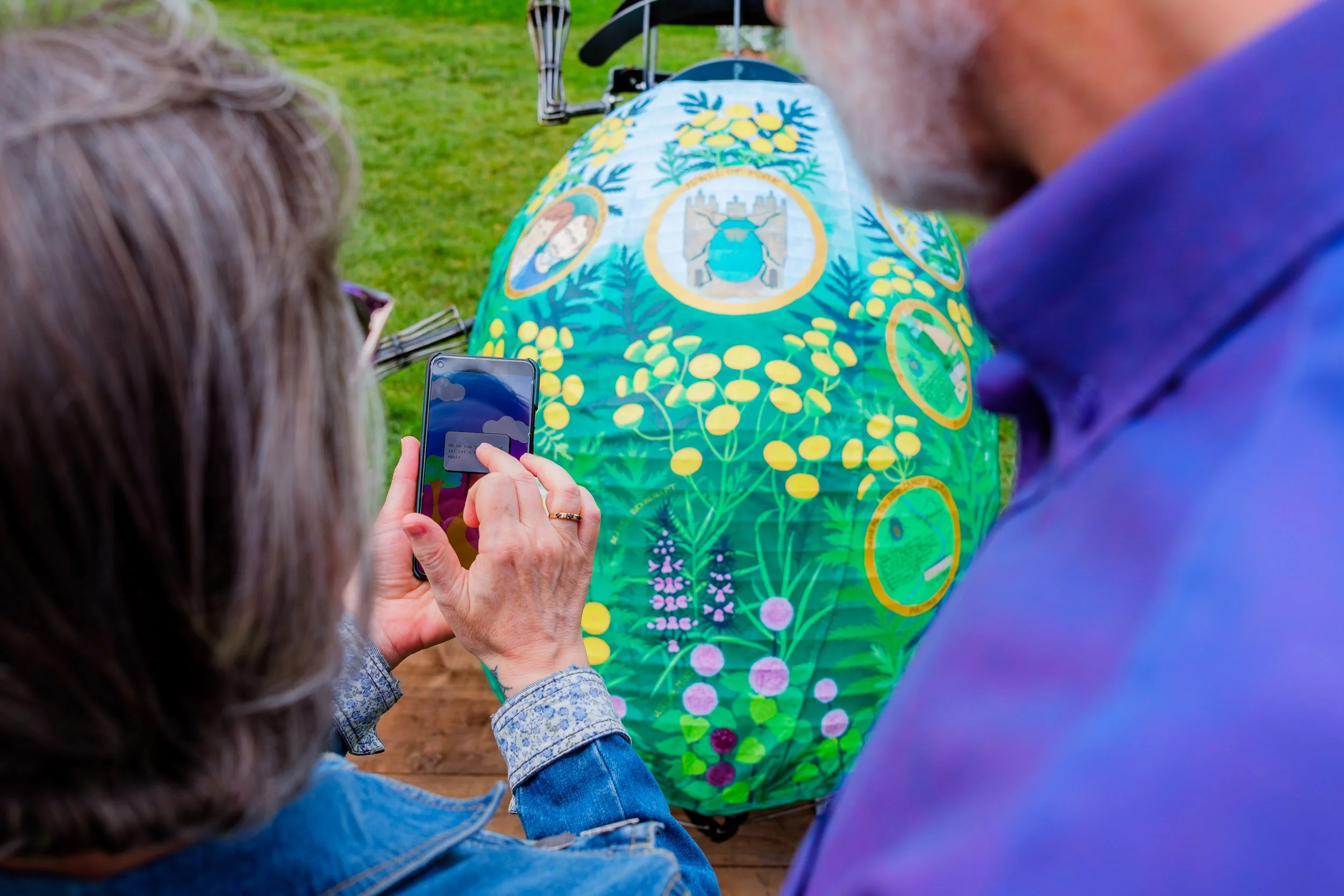 Two people looking at and taking a photo of a large, colorful, painted rock with various painted images and symbols, outdoors on grass.