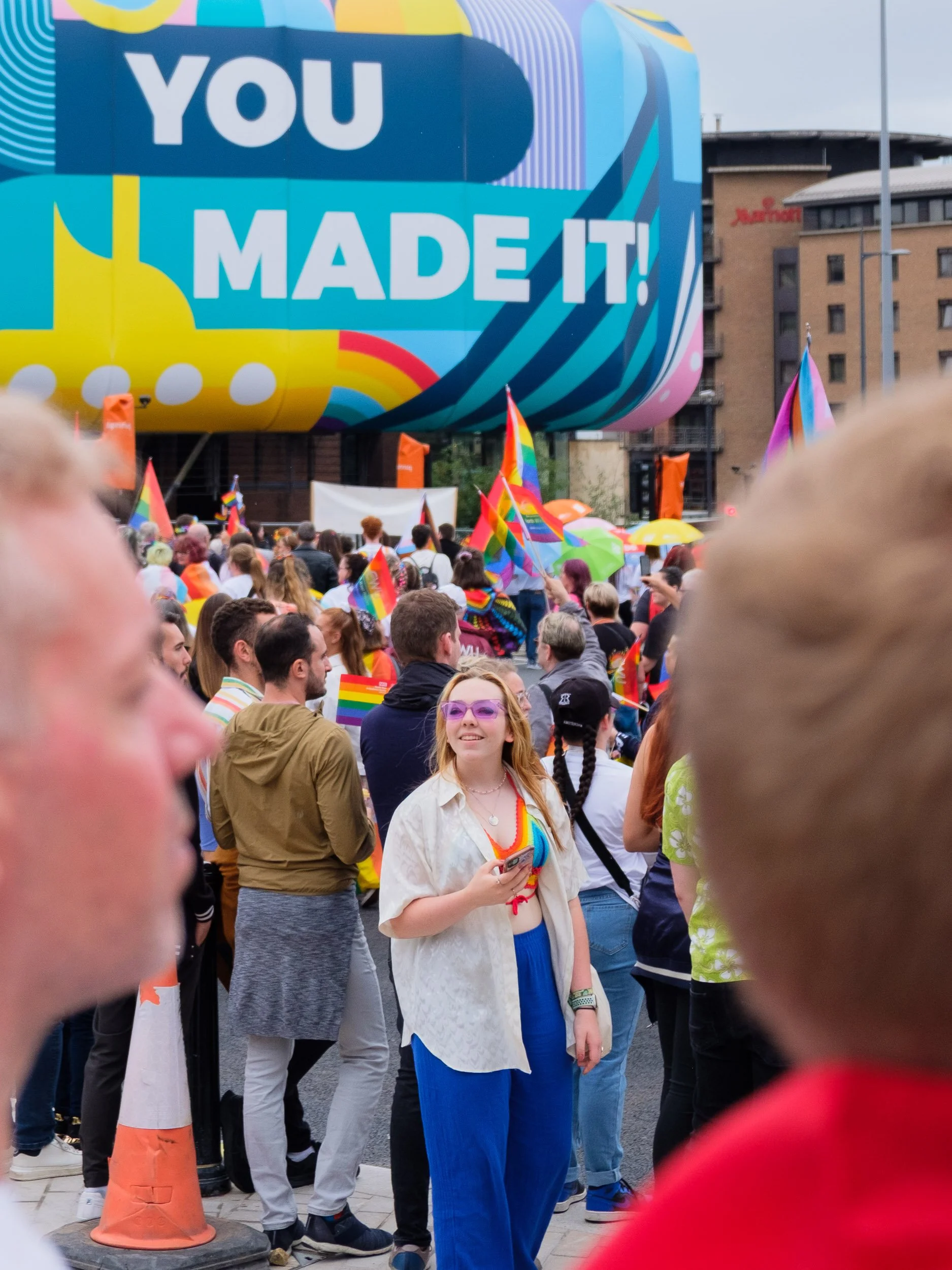Crowd at LGBTQ+ pride event with rainbow flags, large colorful balloon with message "You Made It!" in background