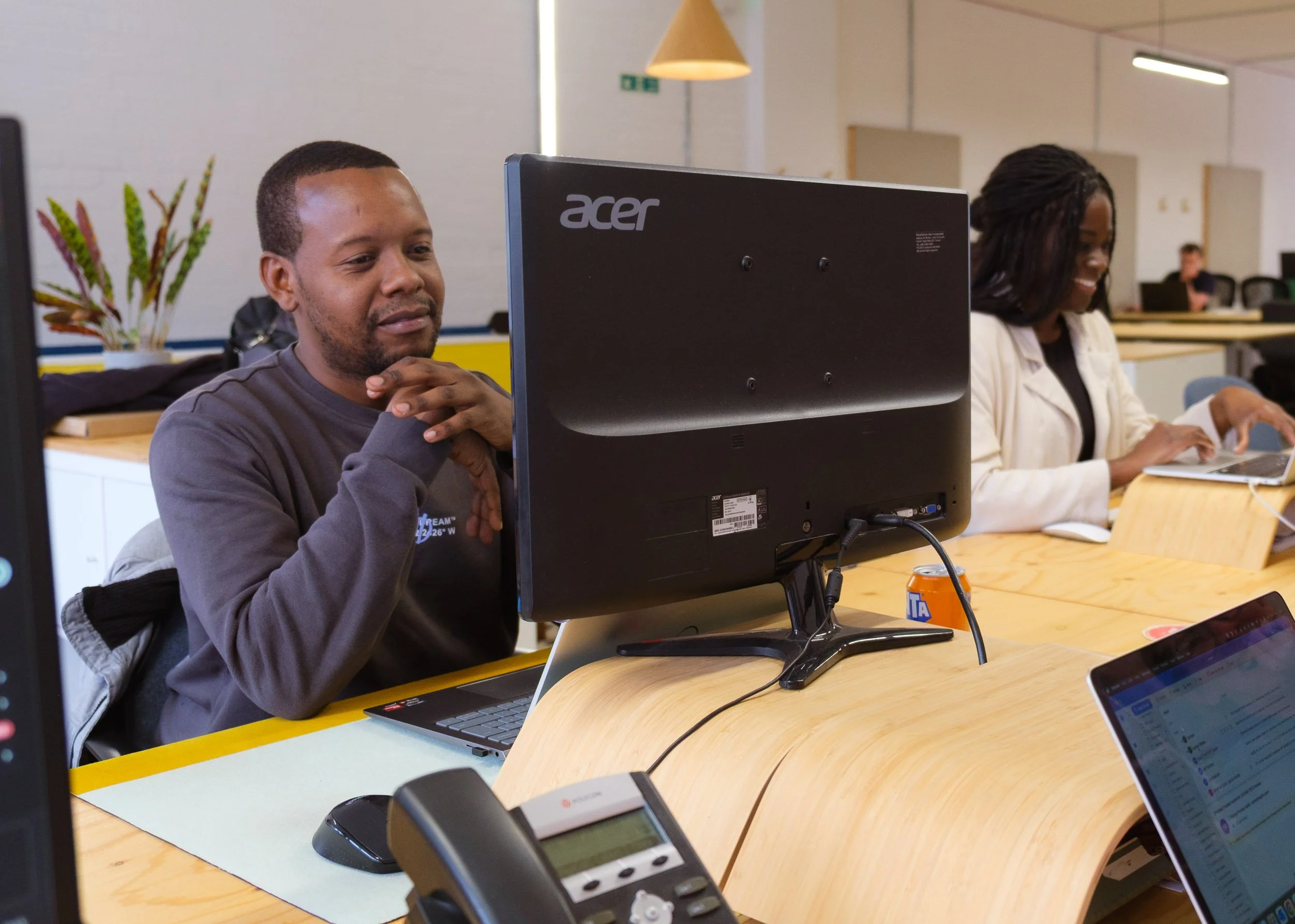 Man in gray sweater sitting at a desk with a large computer monitor, in an office or conference room with other people working, woman in white jacket smiling and working on laptop in background.