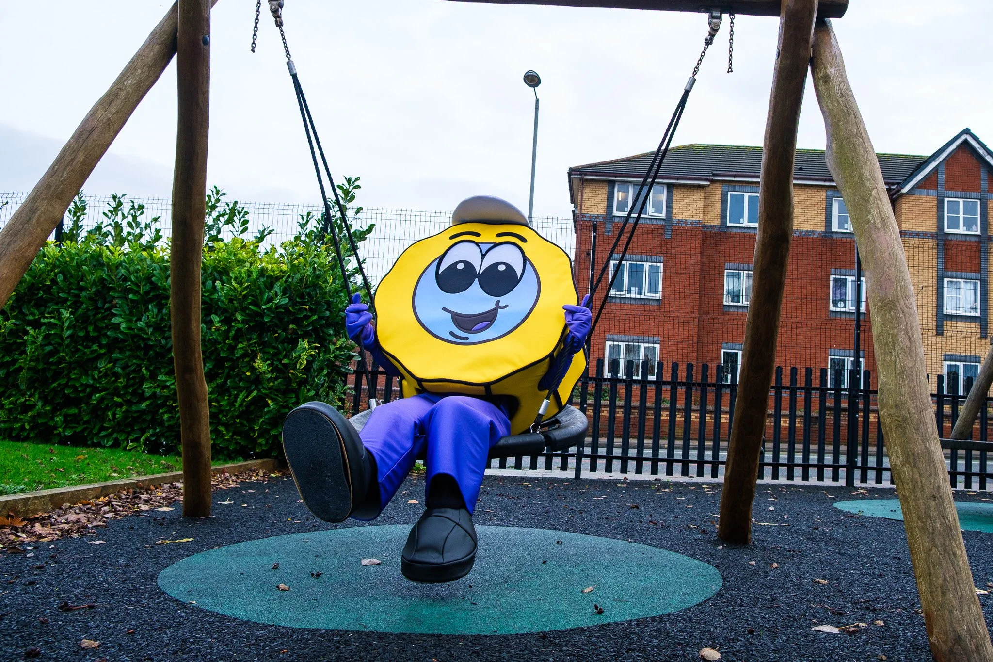 A person dressed in a lemon character costume with a smiley face on the front, sitting on a swing in a playground with a black rubber surface, trees, a fence, and an apartment building in the background.