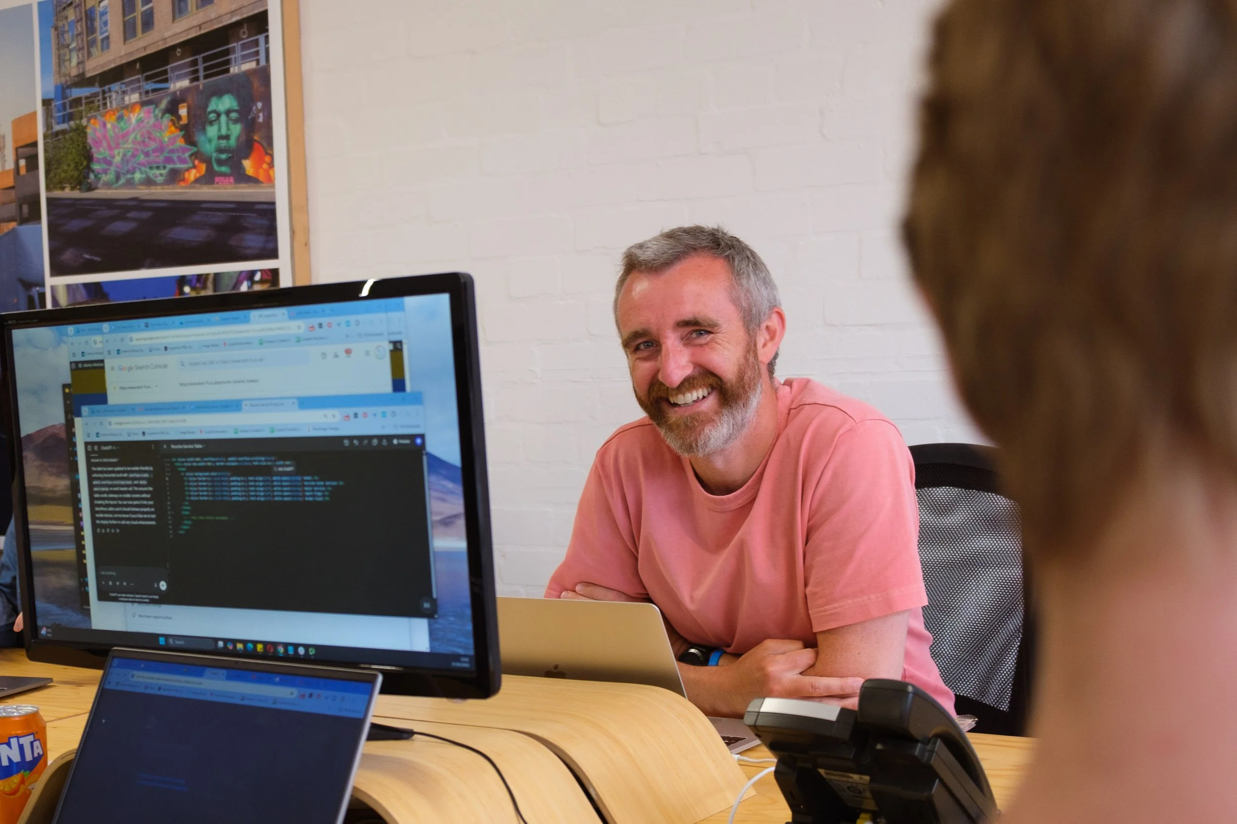 A man with gray hair and a beard is smiling during a meeting, sitting at a desk with computers and a phone. There is a white brick wall and a colorful mural artwork in the background.